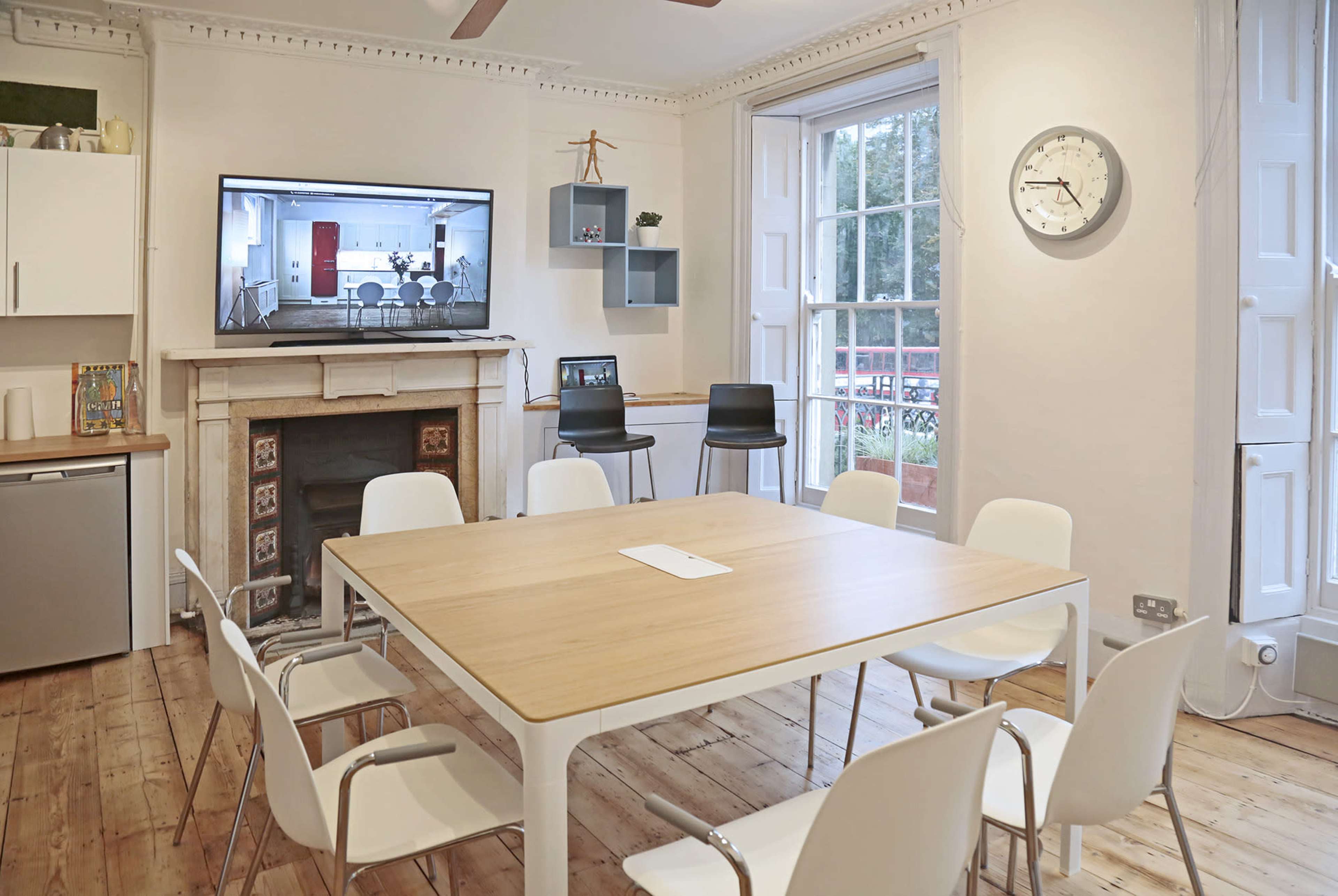 A dining area features a wooden table surrounded by white chairs, a wall-mounted television displaying a meeting room, and large windows with a view.