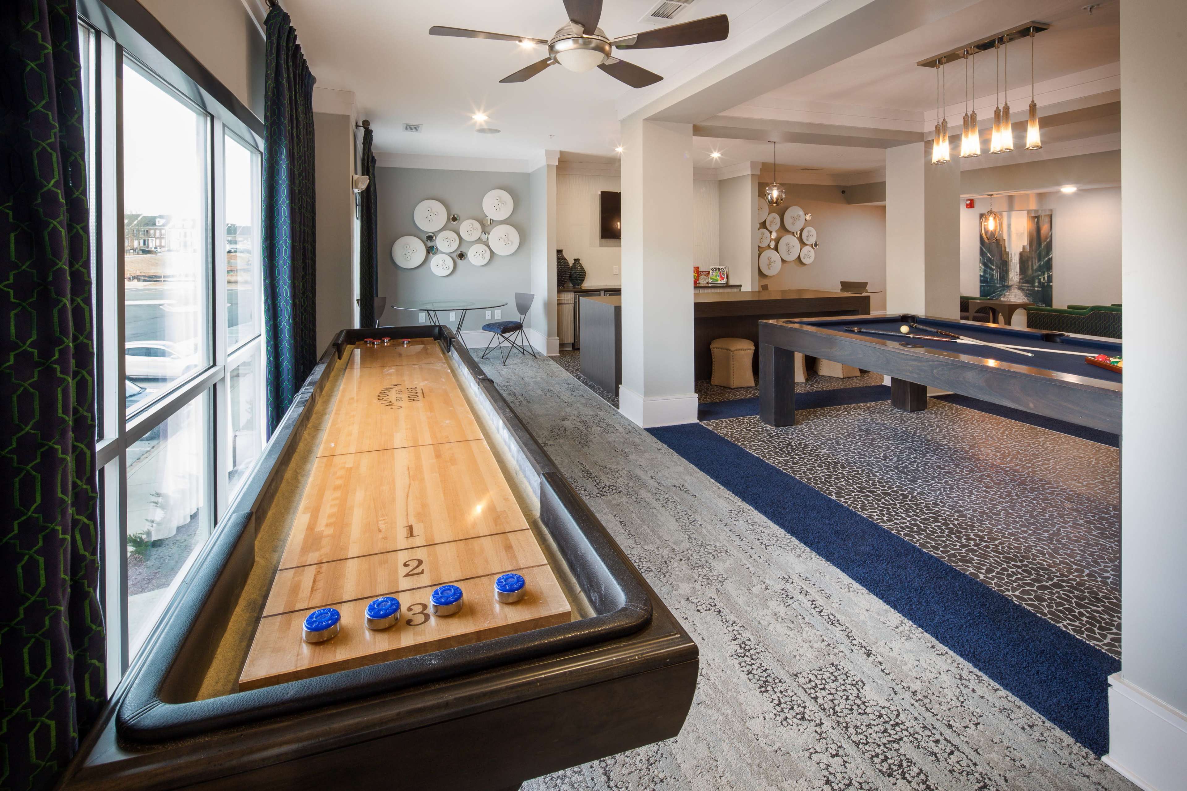The image shows a recreational room featuring a shuffleboard table, a billiards table, and decorative wall plates arranged in a circular pattern.