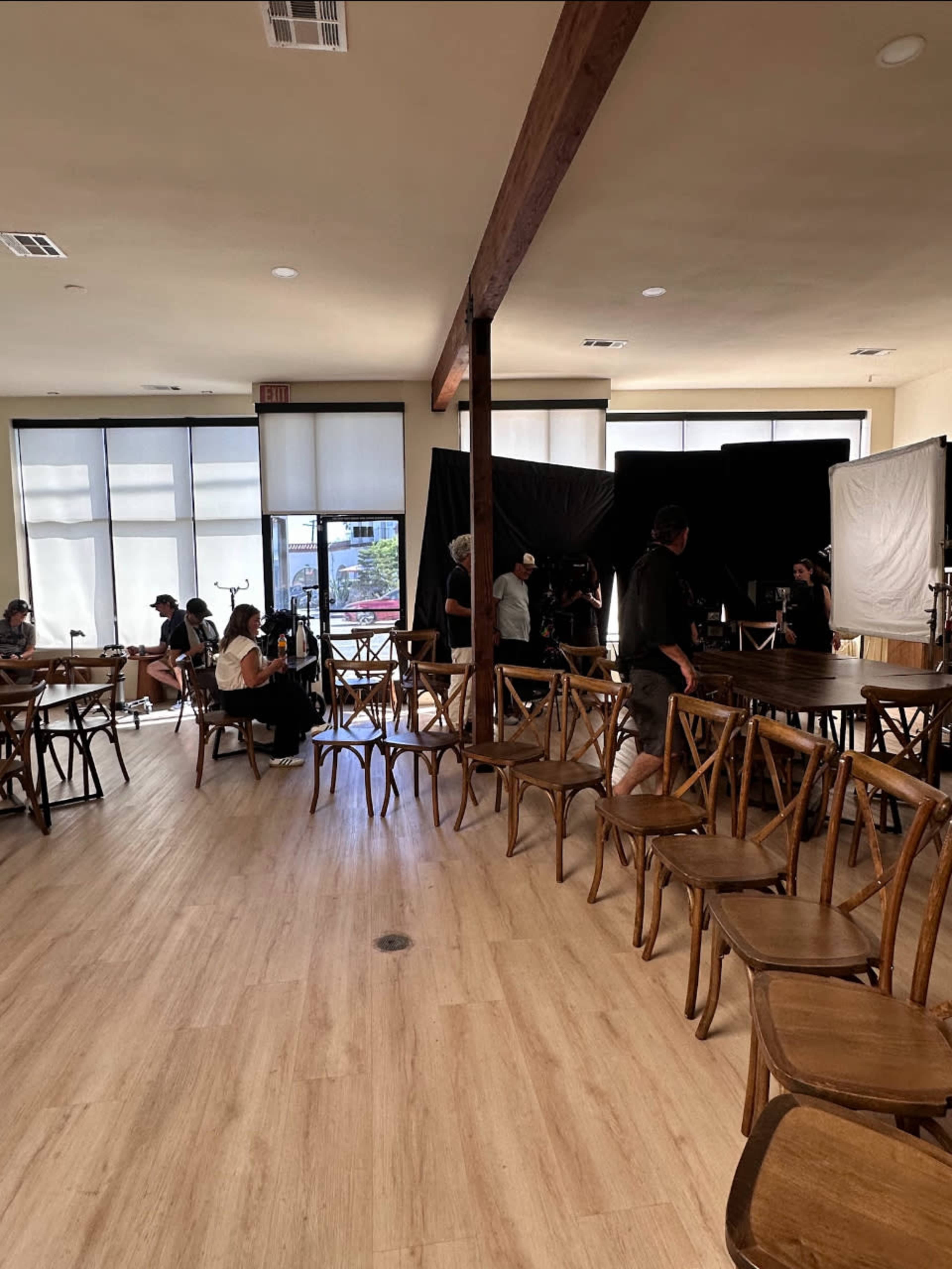 A film crew sets up equipment in a room filled with wooden chairs and large windows.