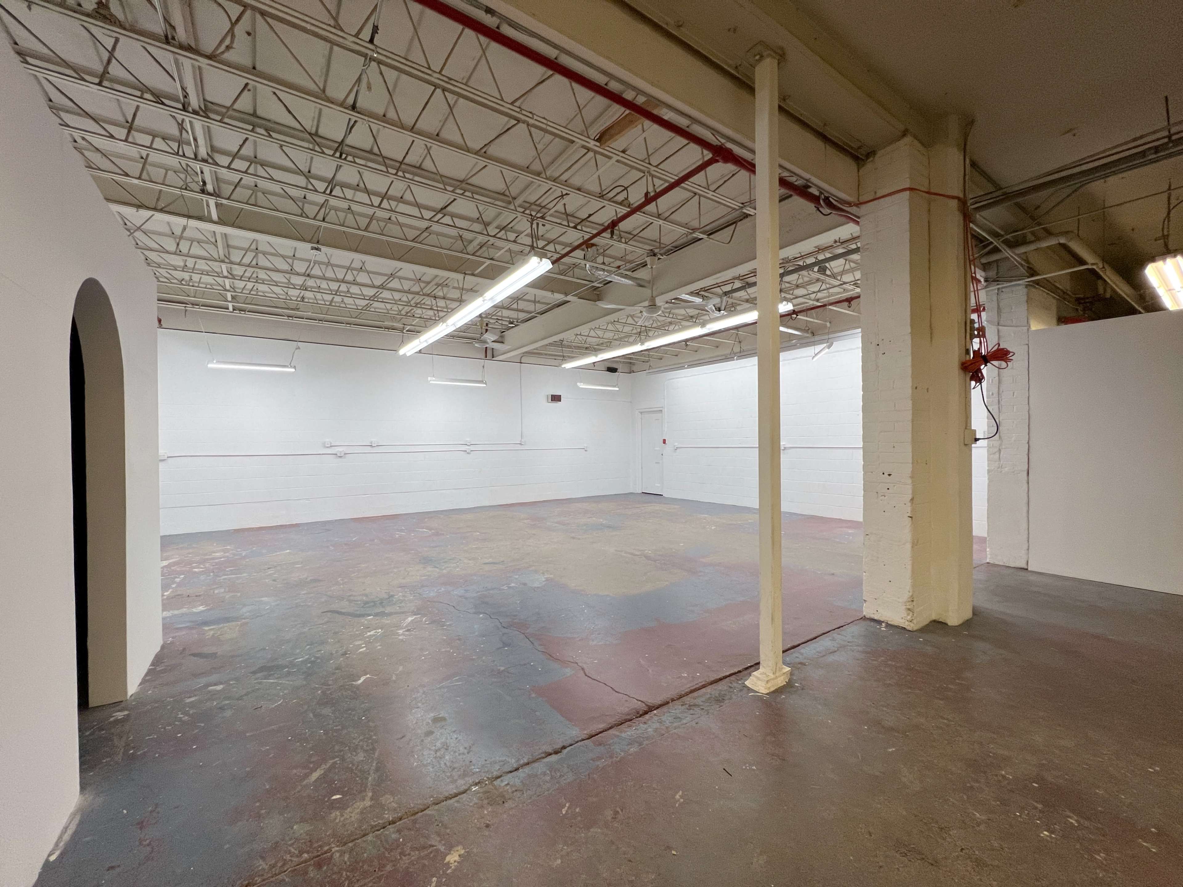 A spacious, empty room with white walls and a concrete floor, featuring exposed pipes and a column in the center.