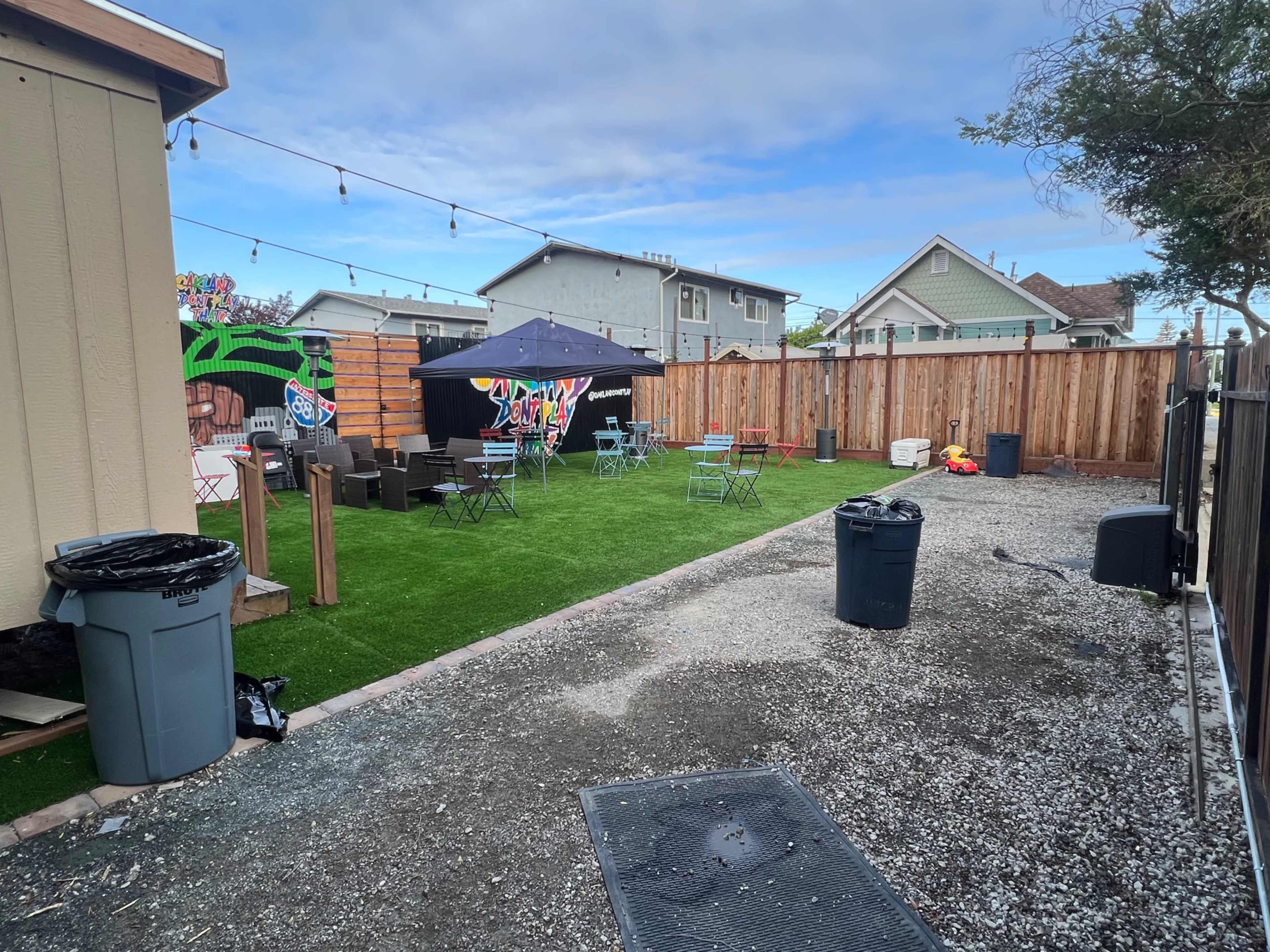 The image shows a backyard area with artificial grass, a tent, various seating arrangements, fences, and trash bins.