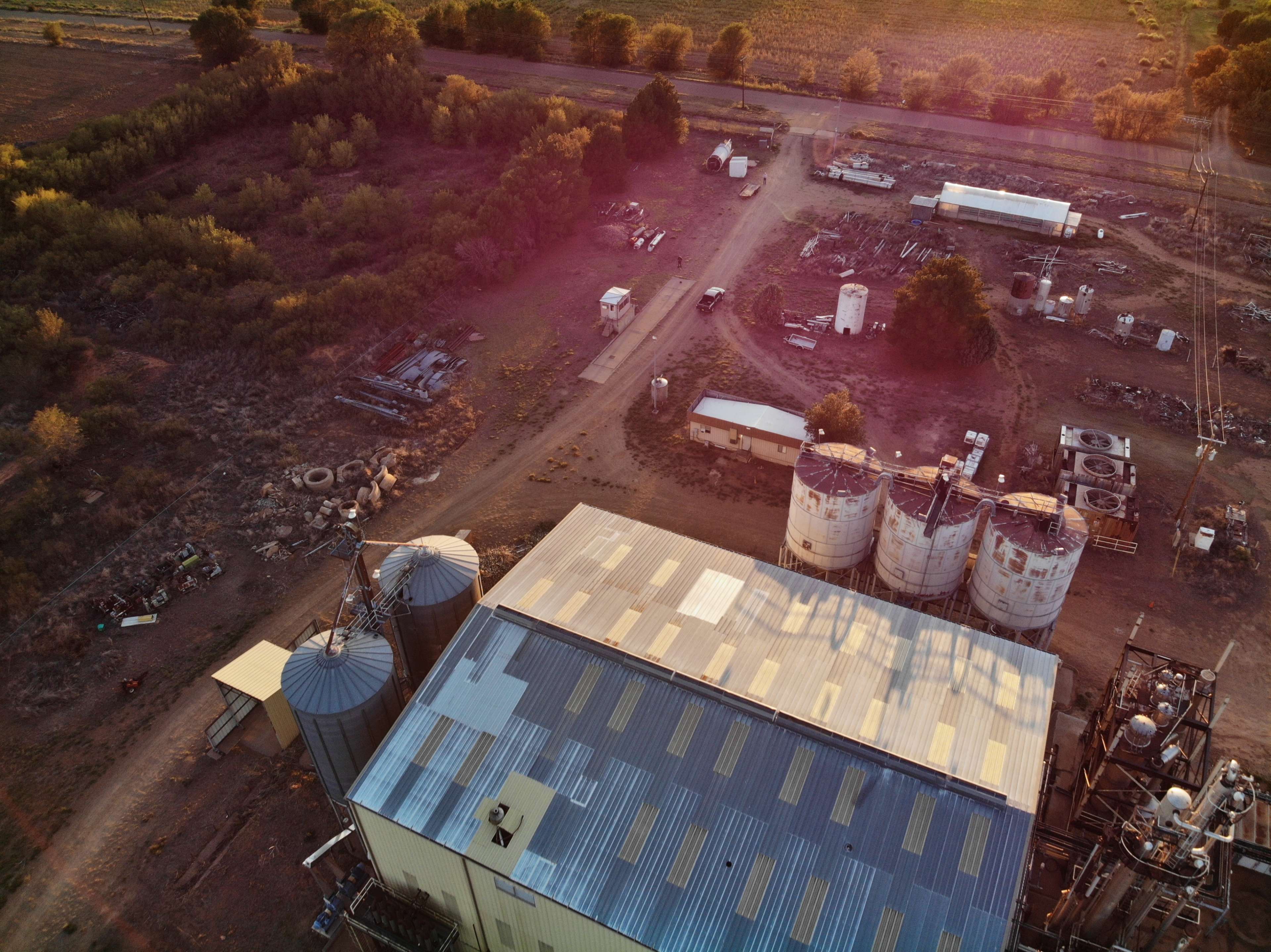 An aerial view shows agricultural silos, machinery, and buildings surrounding a dirt road in a rural setting.
