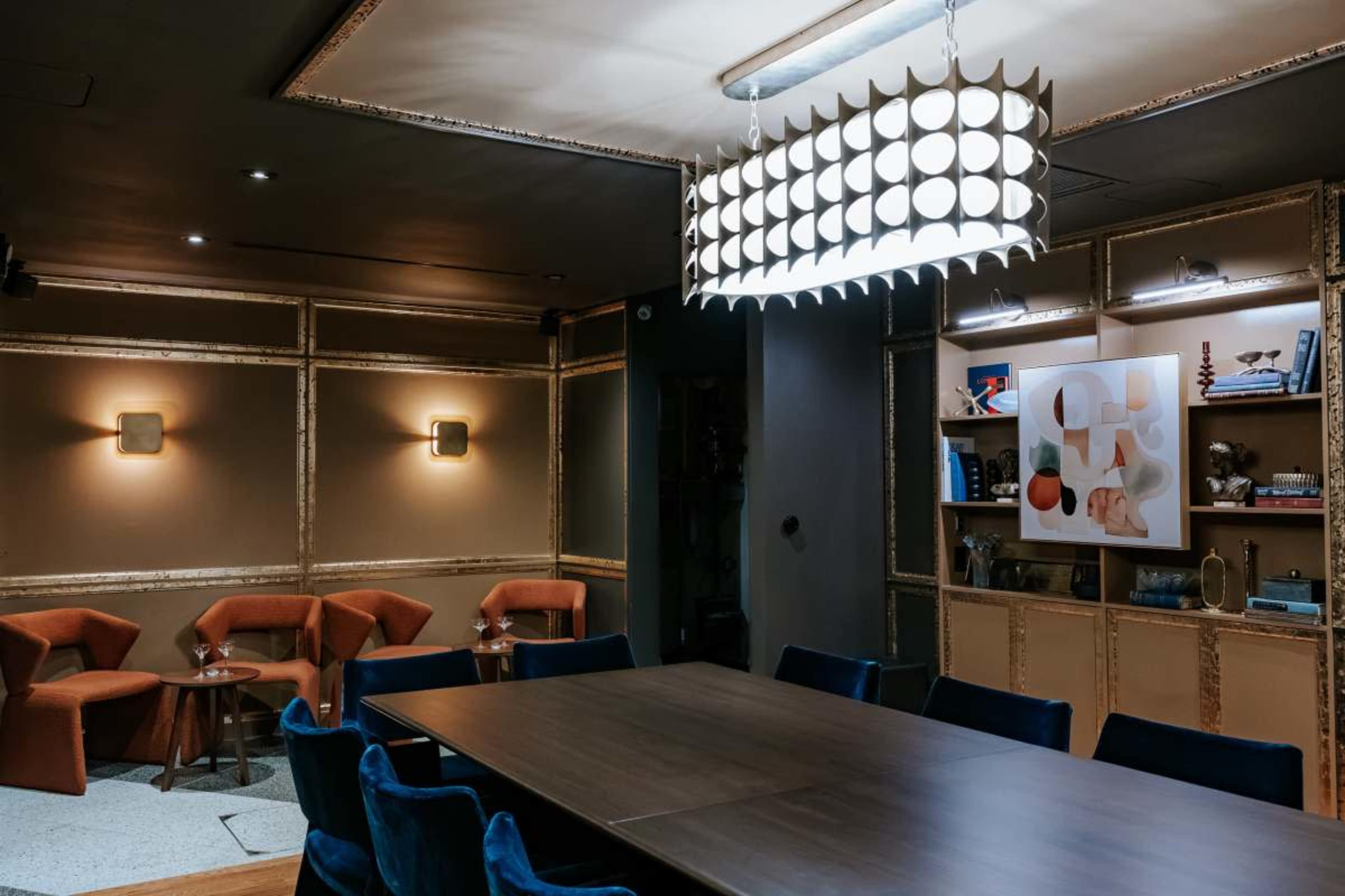 A stylish meeting room features a long wooden table surrounded by plush blue and orange chairs, with a modern chandelier and decorative shelving in the background.