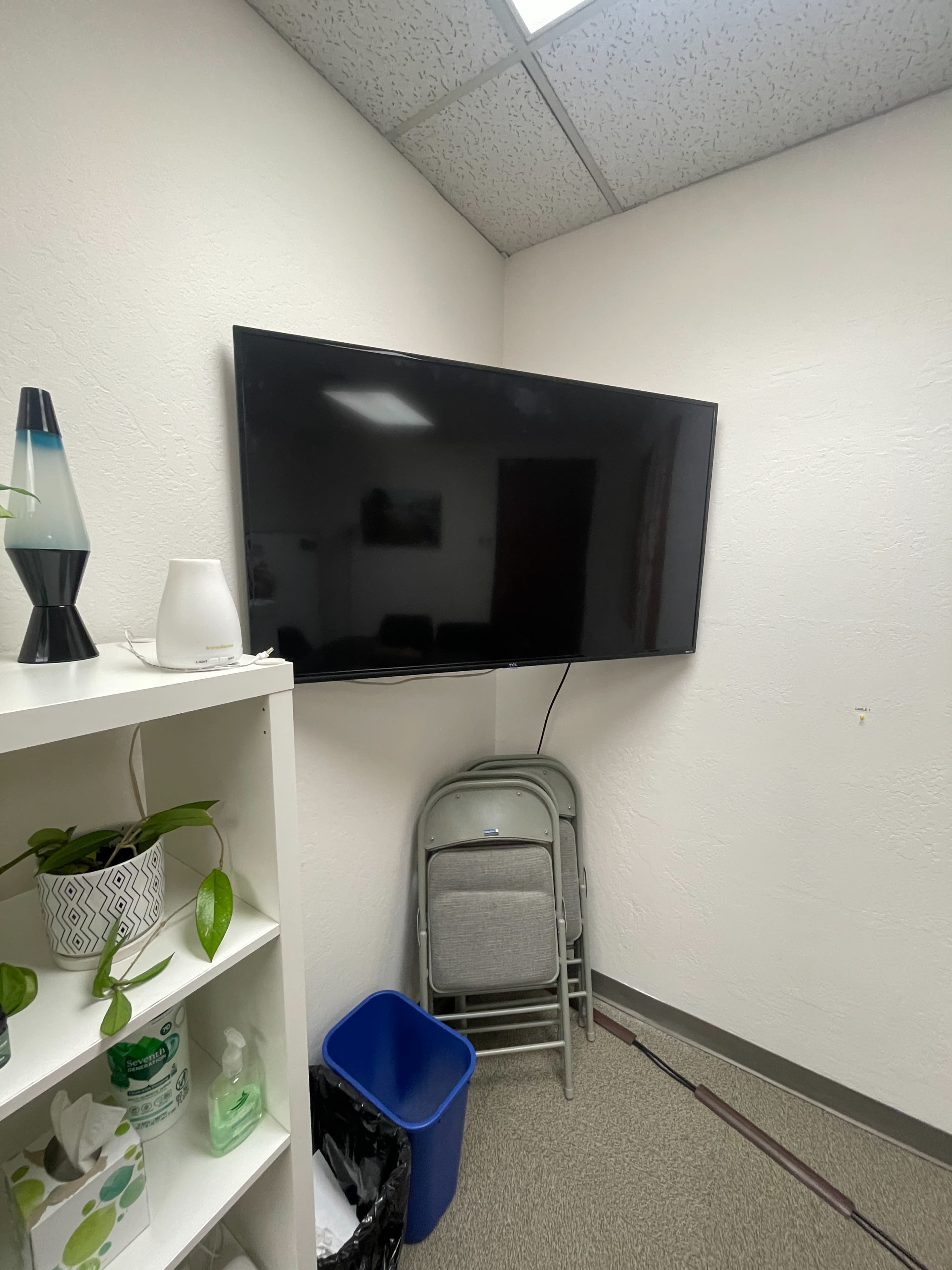A flat-screen TV mounted on a wall in a small room, with a folding chair and a blue recycling bin positioned on the floor below it.
