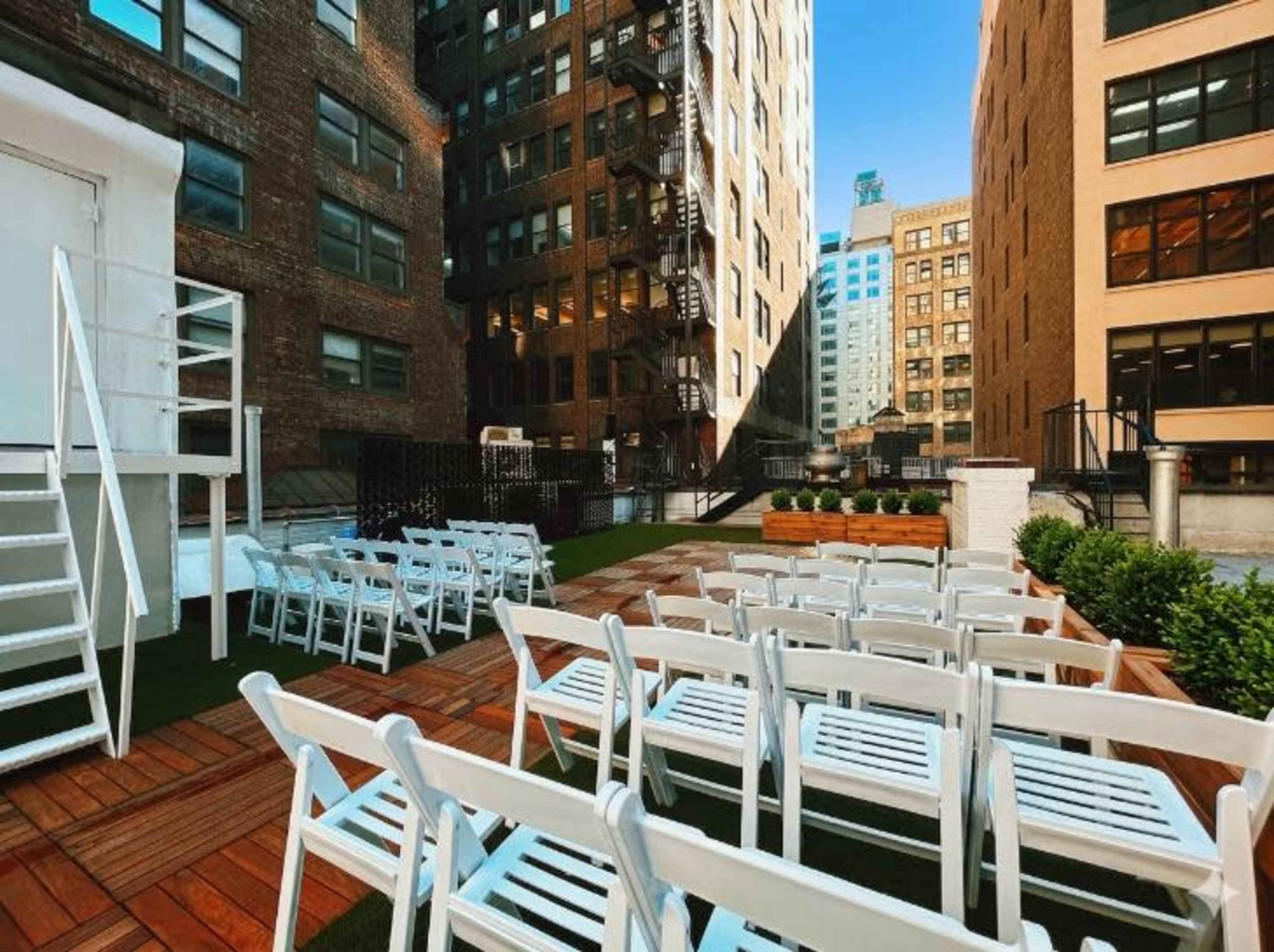 The image shows an outdoor event space with white folding chairs arranged in rows on a wooden platform surrounded by tall buildings.