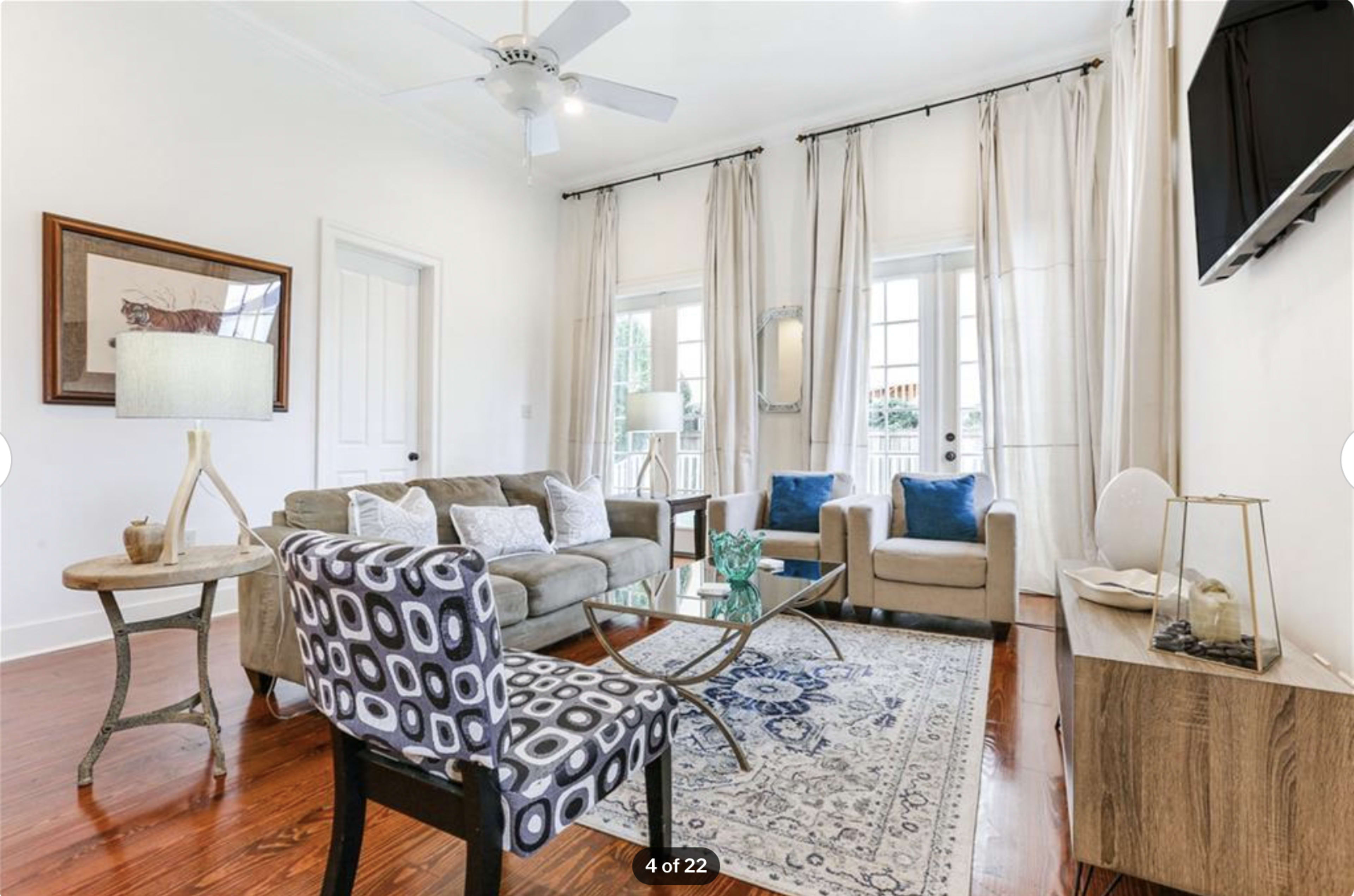A bright living room with a couch, two armchairs, a glass coffee table, and large windows draped with light curtains.