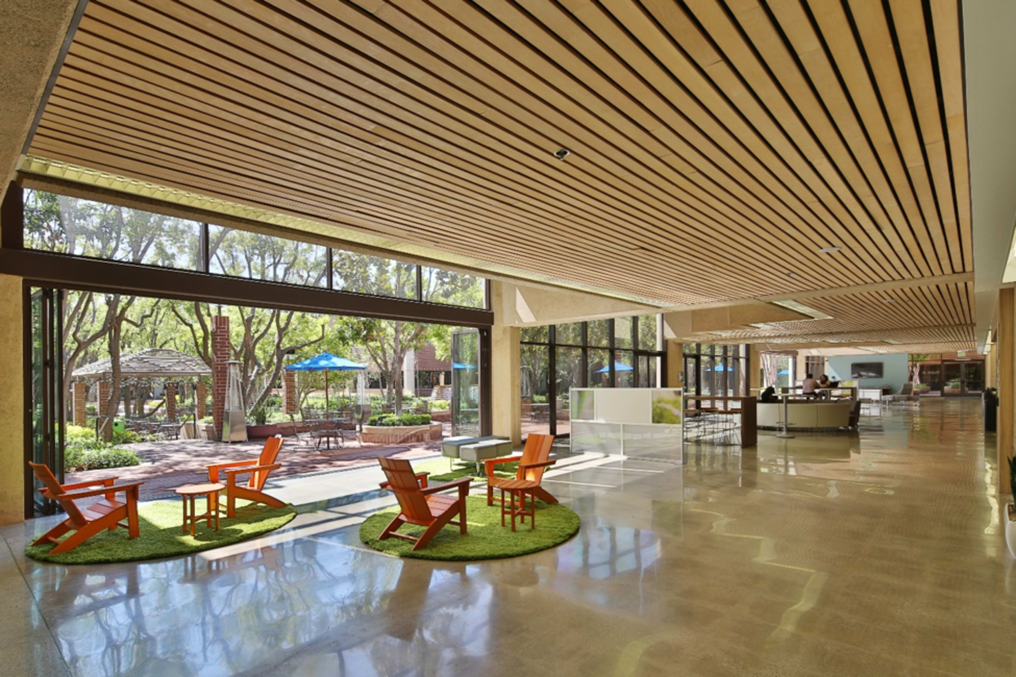 A modern interior space featuring wooden beams, a smooth floor, and orange chairs arranged on green rugs, with large windows offering views of an outdoor area with trees and seating.