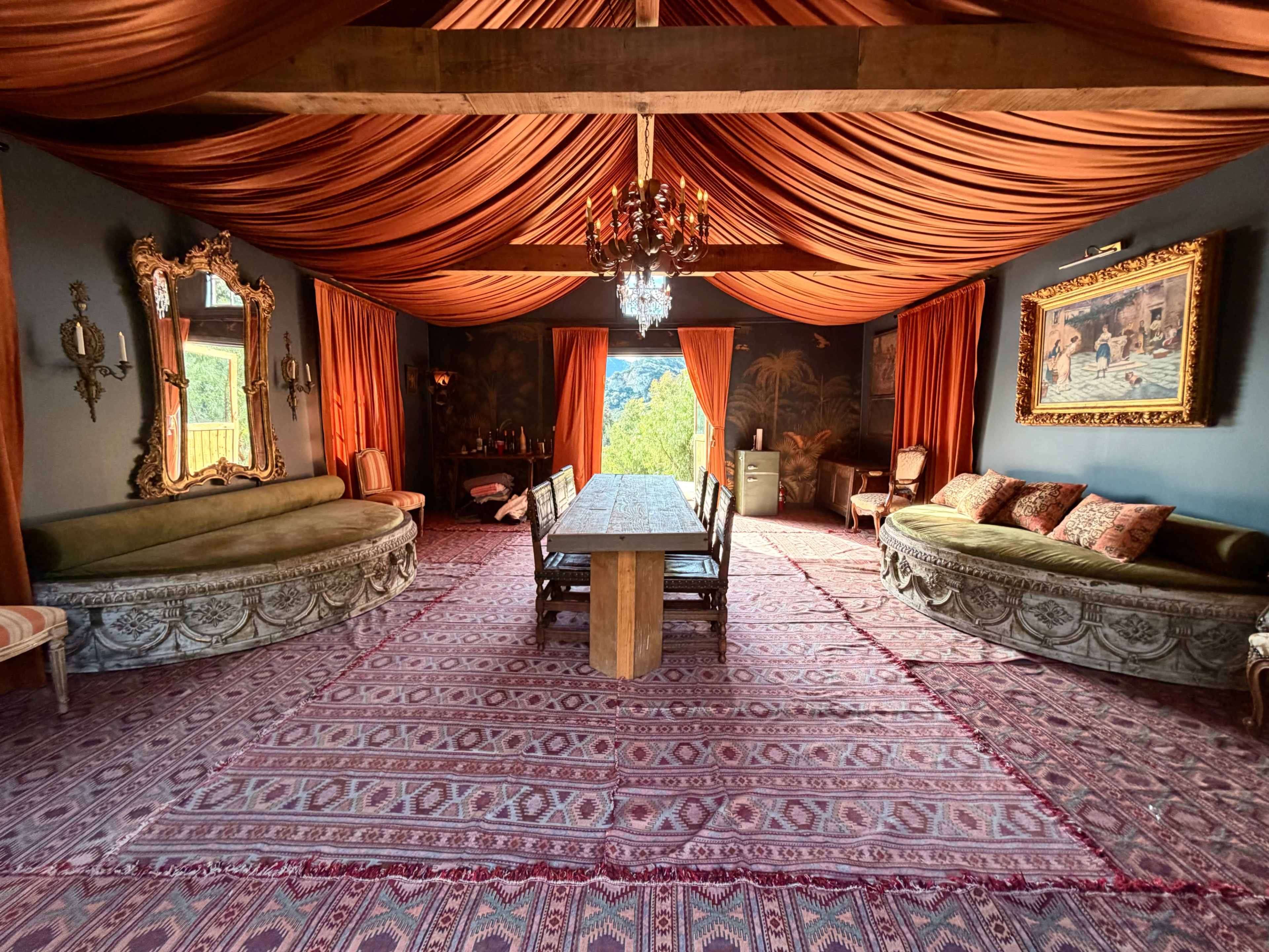 The room features a long wooden dining table surrounded by chairs, with ornate sofas and rich drapery overhead.