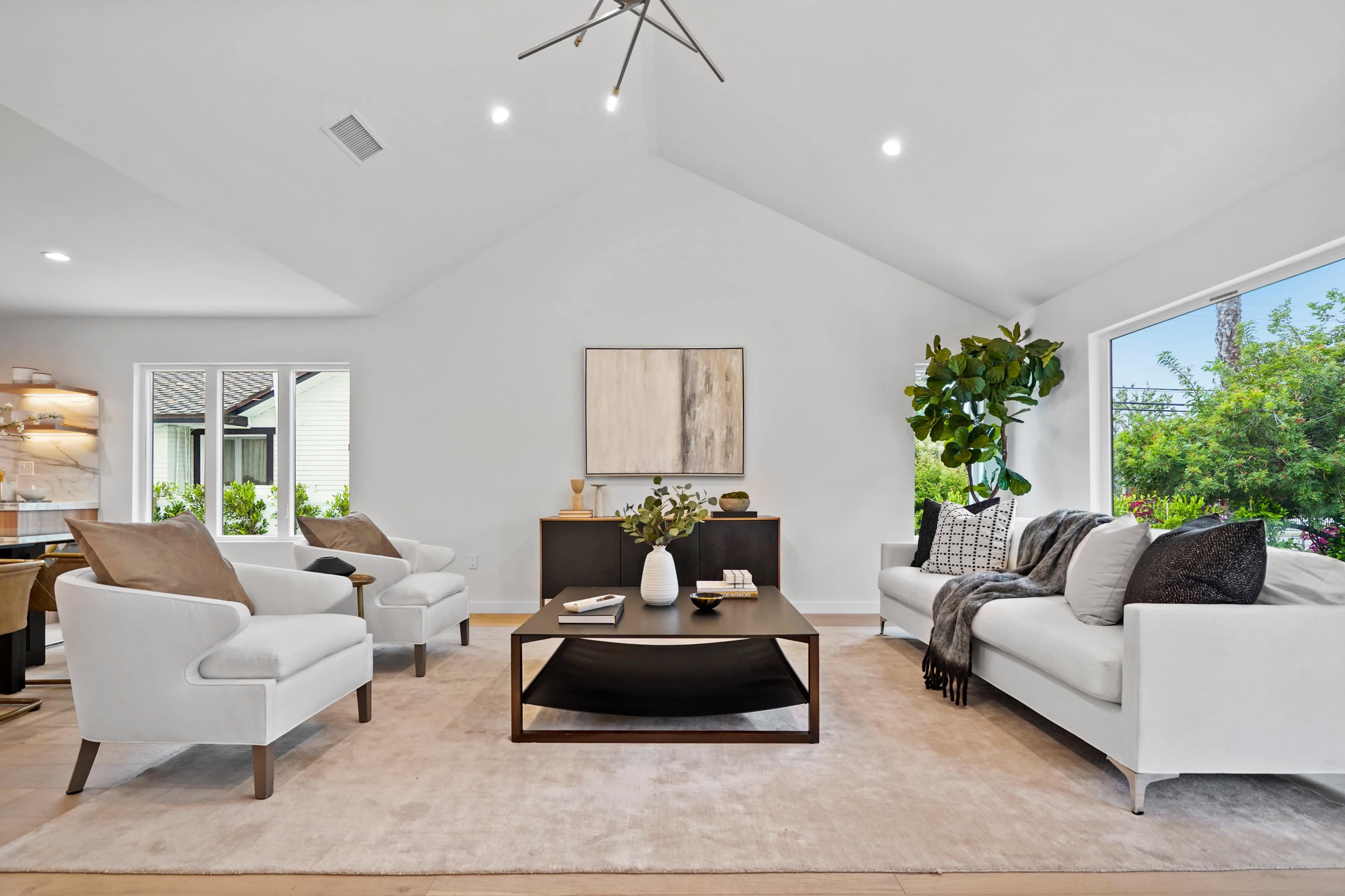A modern living room features a black coffee table at the center, surrounded by two white armchairs and a white sofa against neutral walls and large windows.