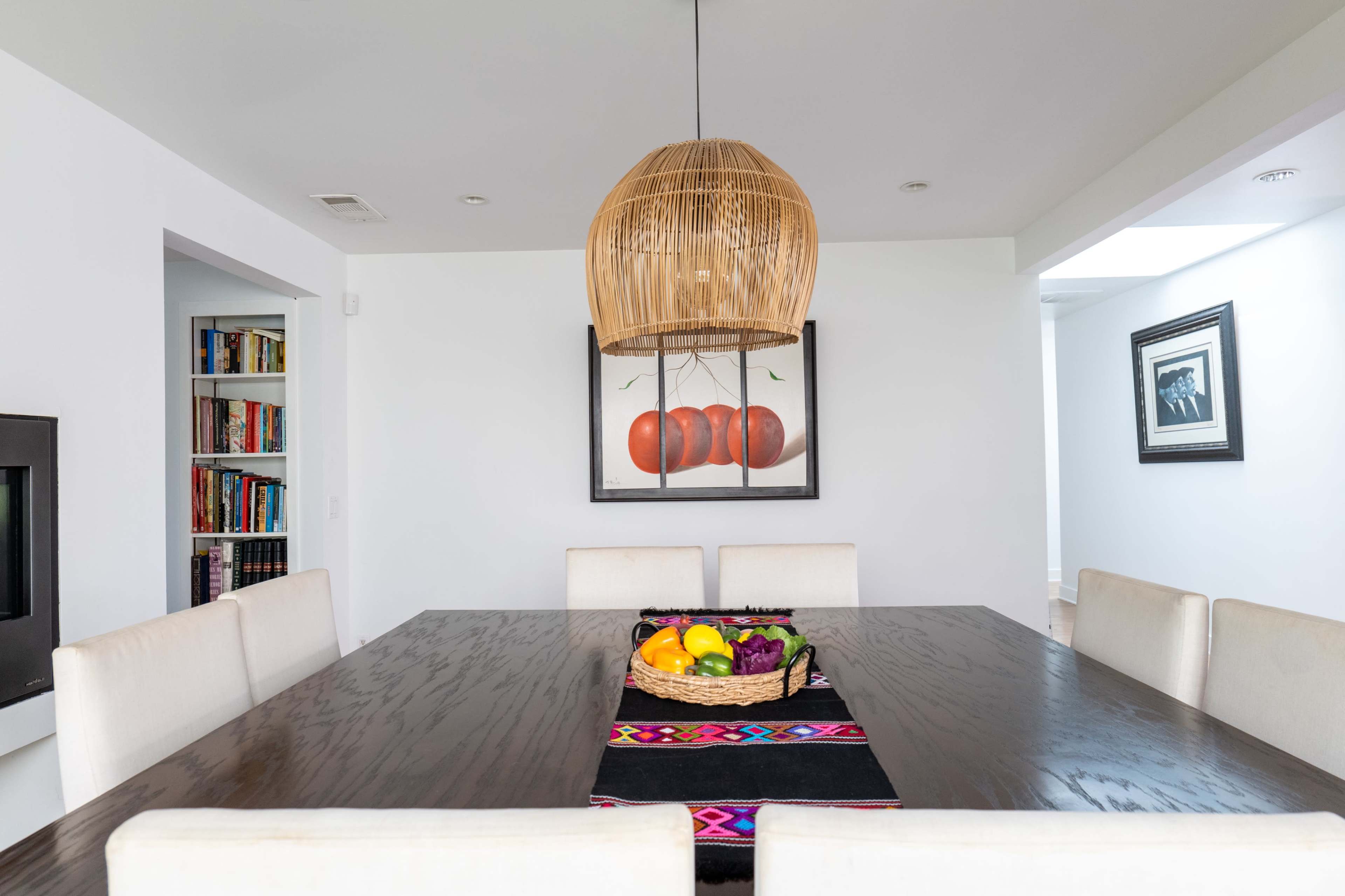 A spacious dining area features a large wooden table with a fruit basket at its center, surrounded by beige chairs and illuminated by a woven pendant lamp.