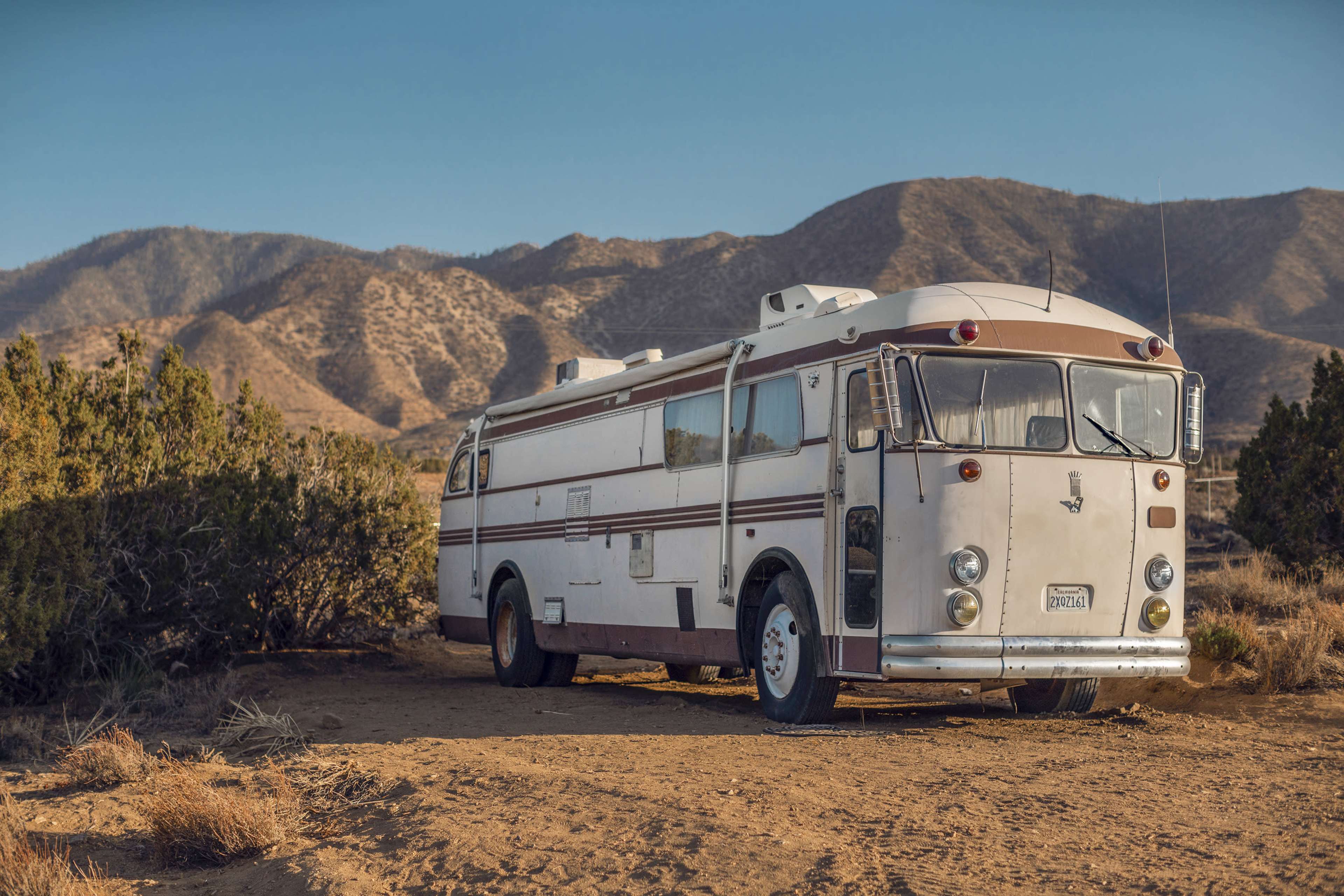 An old bus is parked on a dirt path surrounded by sparse vegetation with mountains in the background.