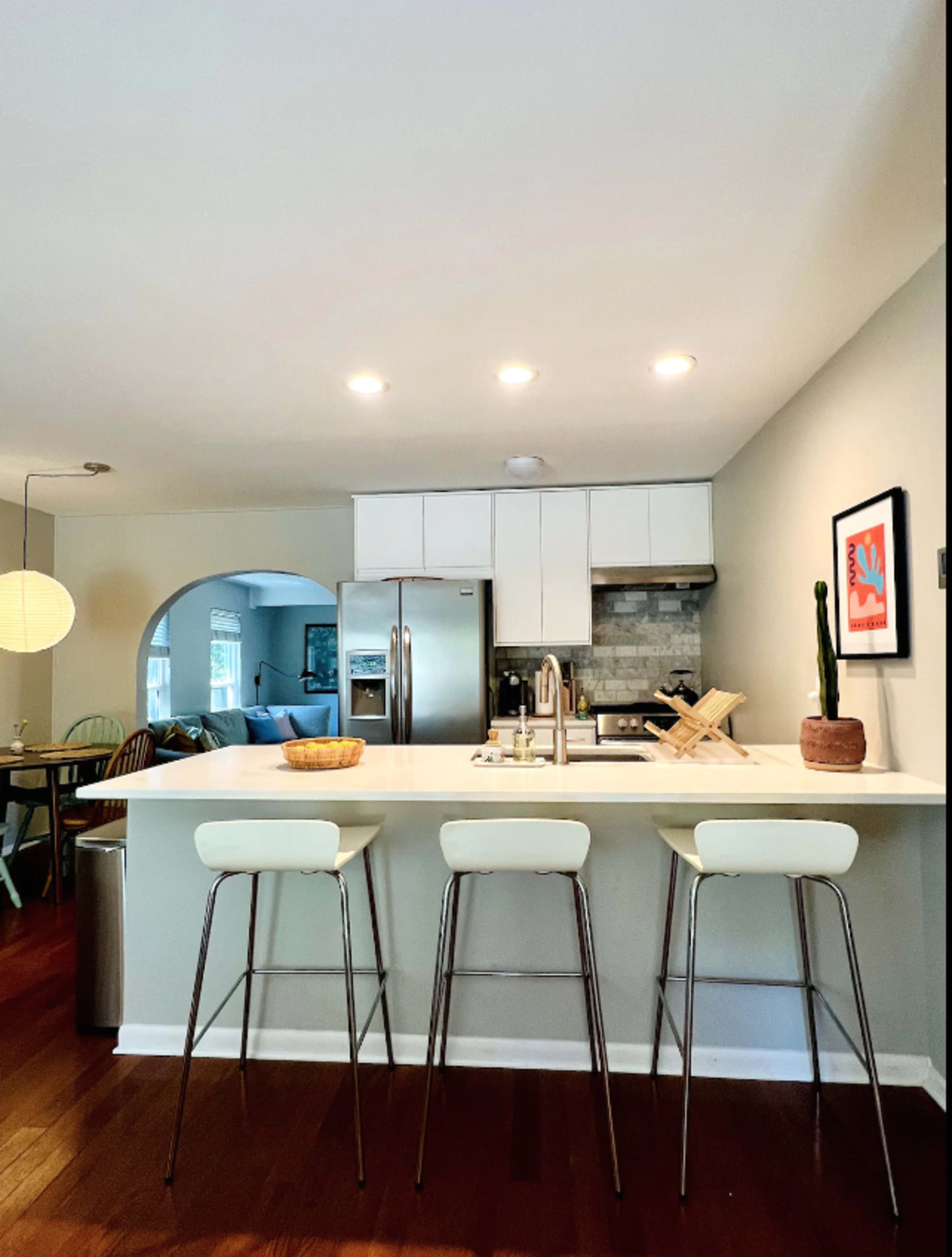 A modern kitchen features a light countertop with three high stools, stainless steel appliances, and an open layout leading to a living area.