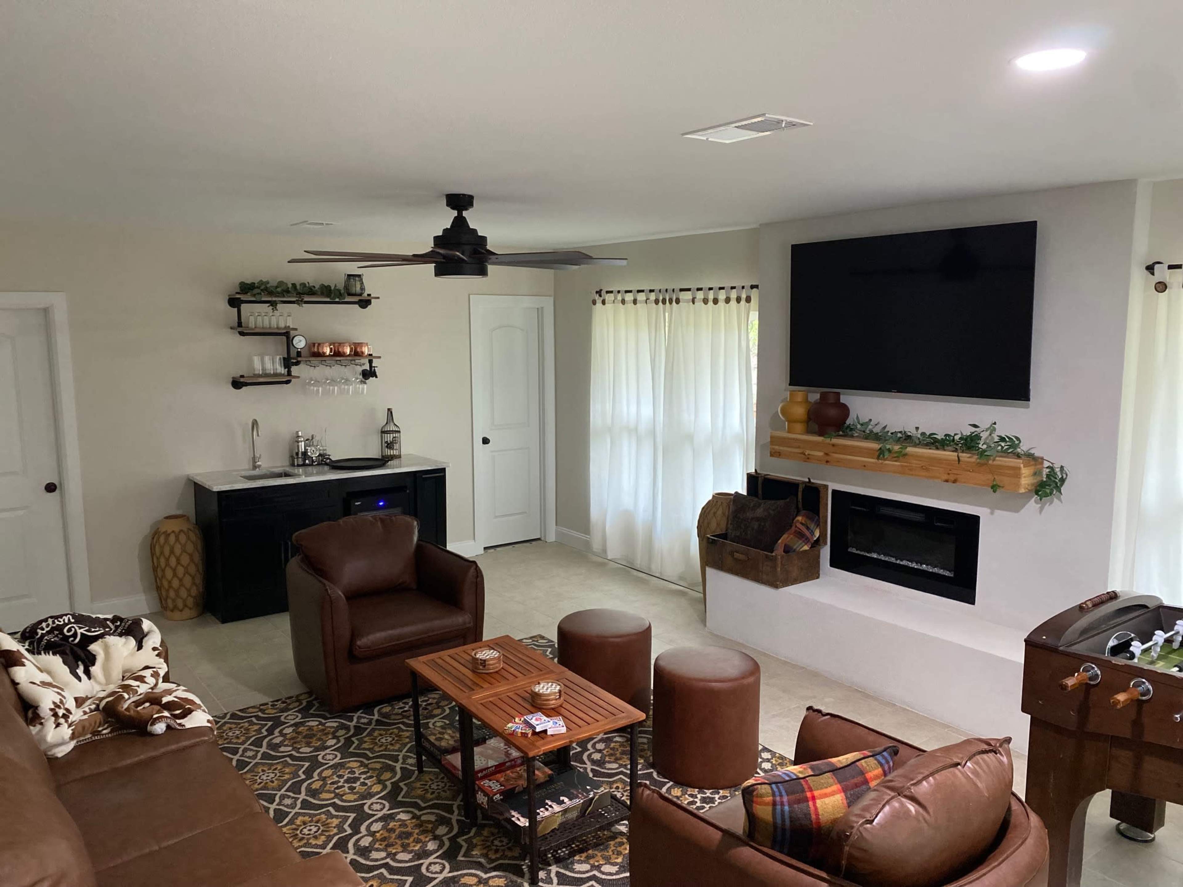 A cozy living room featuring a seating area with brown leather chairs, a coffee table, and a mounted TV over a fireplace, along with a small bar area.