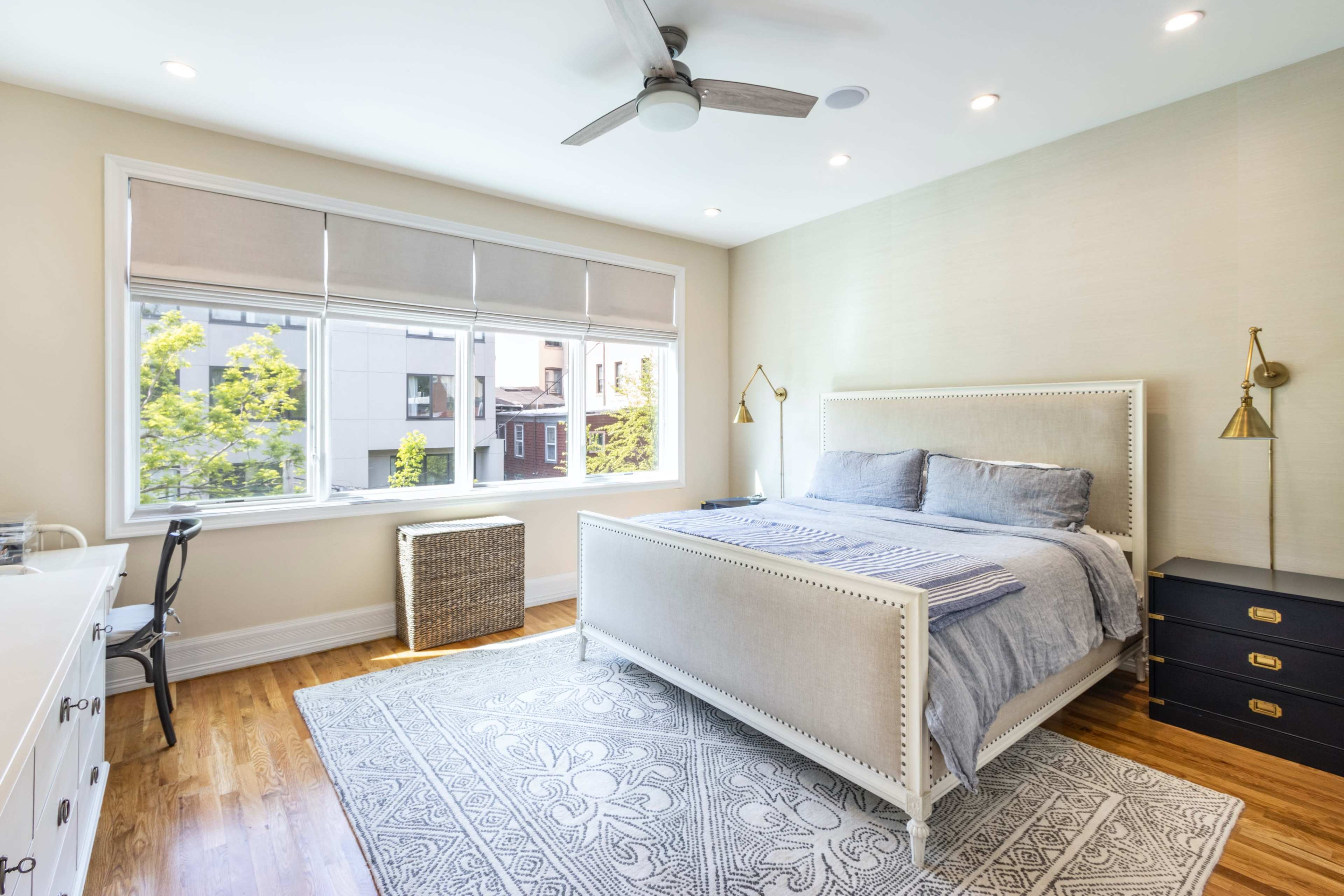 A bright bedroom features a large bed with gray bedding, a window with natural light, a wooden floor, and a desk against a wall.
