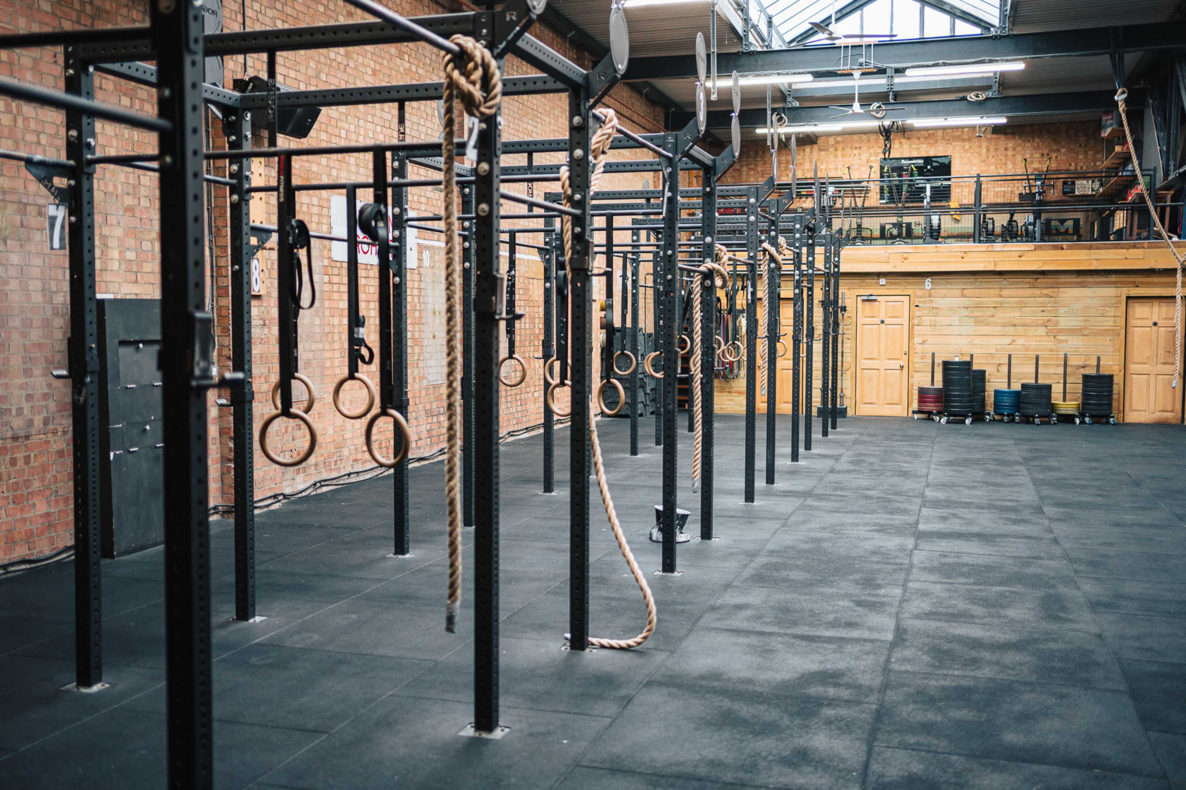 The image shows a spacious gym with pull-up bars and gymnastic rings hanging from the ceiling, set on a rubber floor surface.