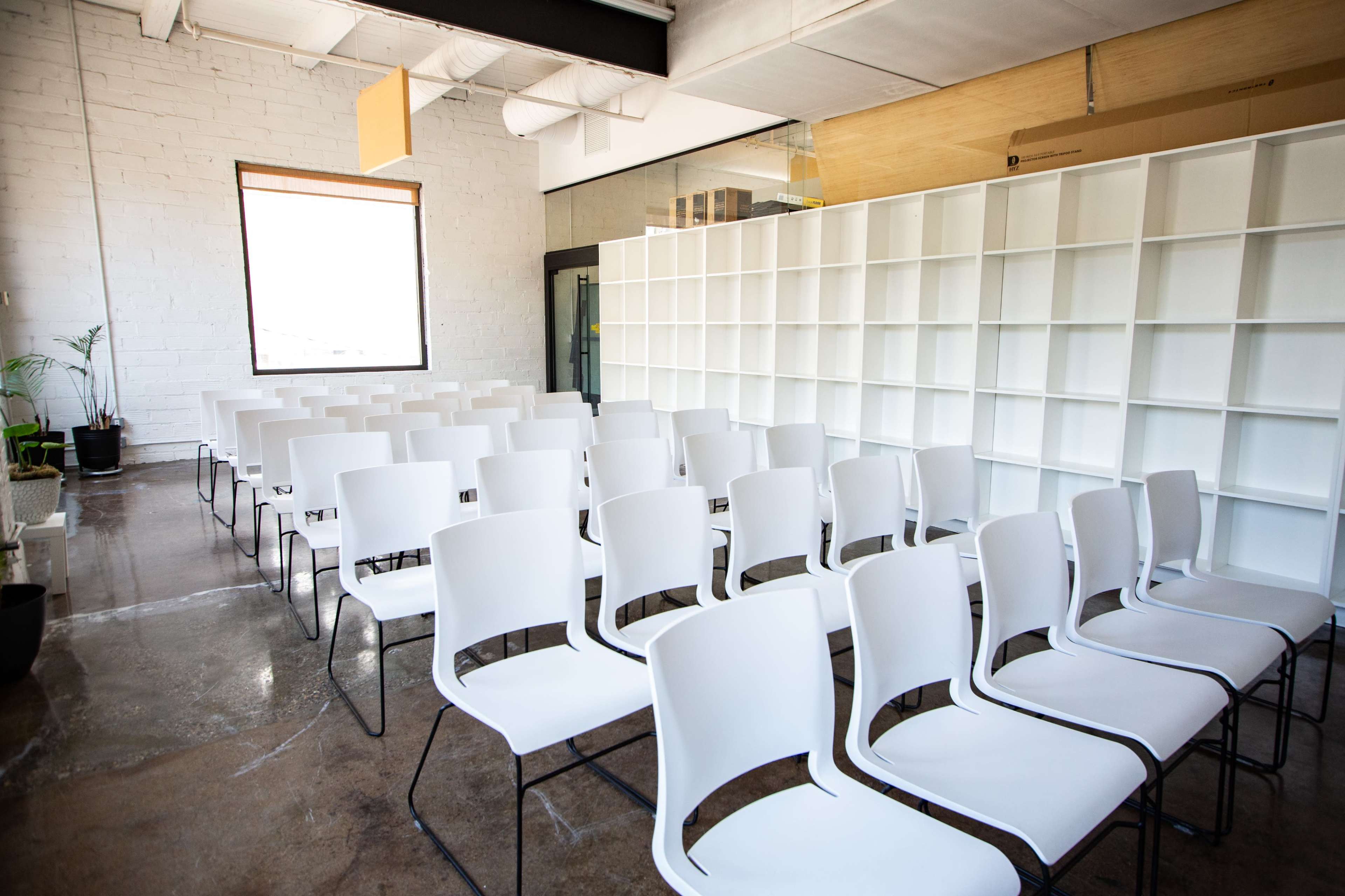 A spacious room features neat rows of white chairs facing a large window and a white shelving unit.