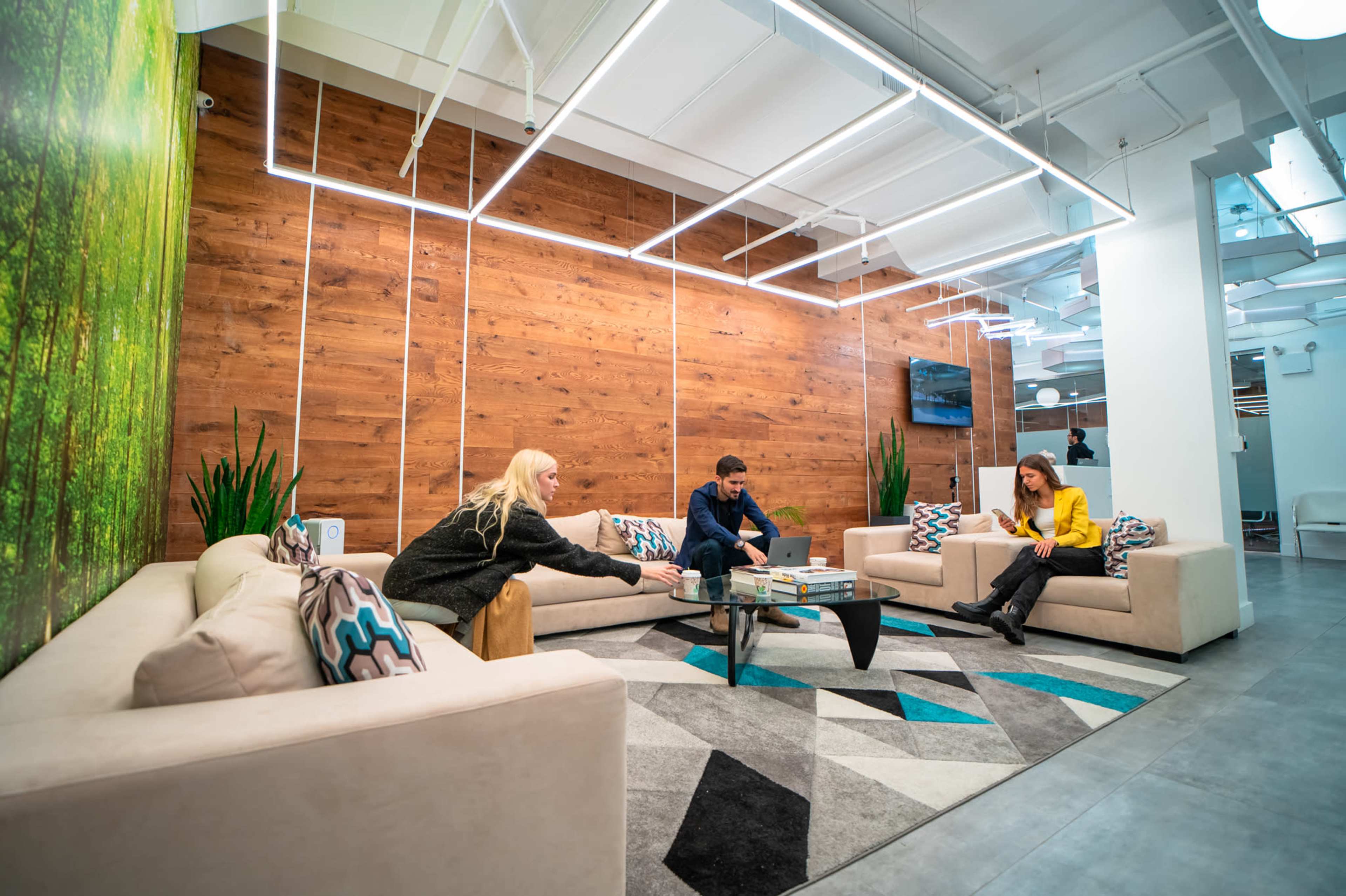 A modern office lounge features three people engaged in conversation around a coffee table, with a backdrop of green foliage and wooden wall panels.