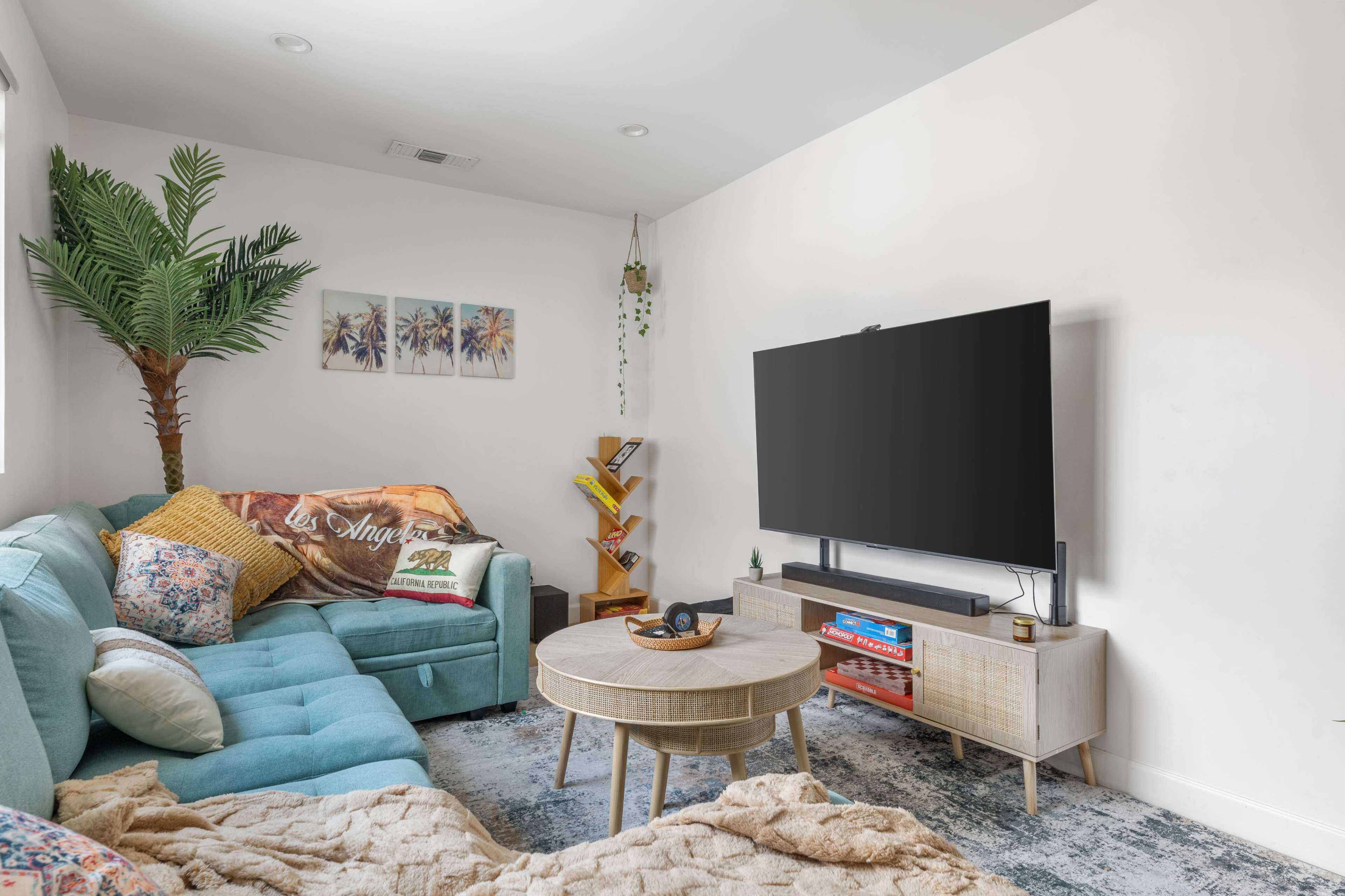 The image shows a cozy living room featuring a blue sectional sofa, a wooden coffee table, a television mounted on the wall, and decorative plants.