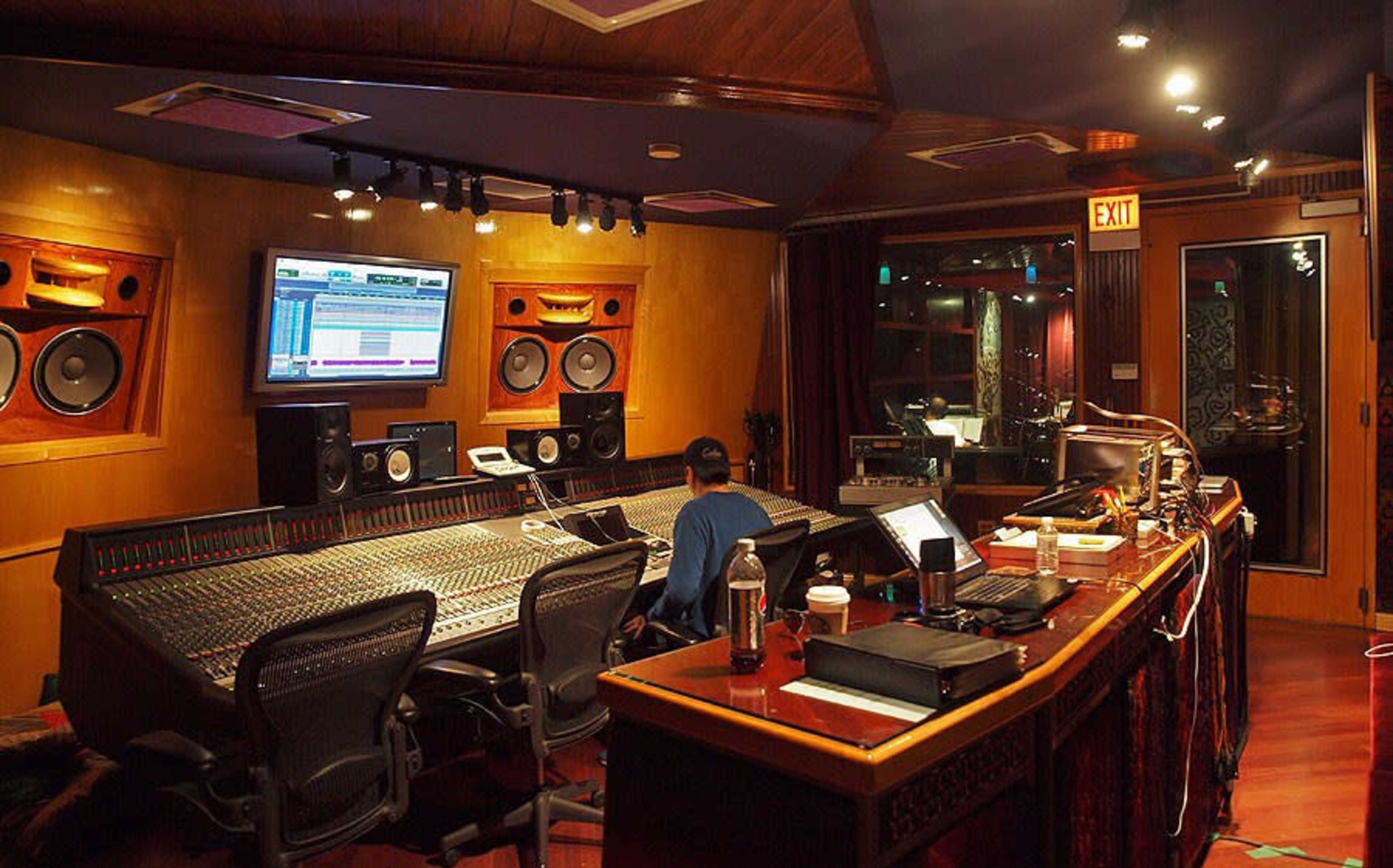 A person is seated at a large mixing console in a dimly lit recording studio equipped with speakers and computer screens.