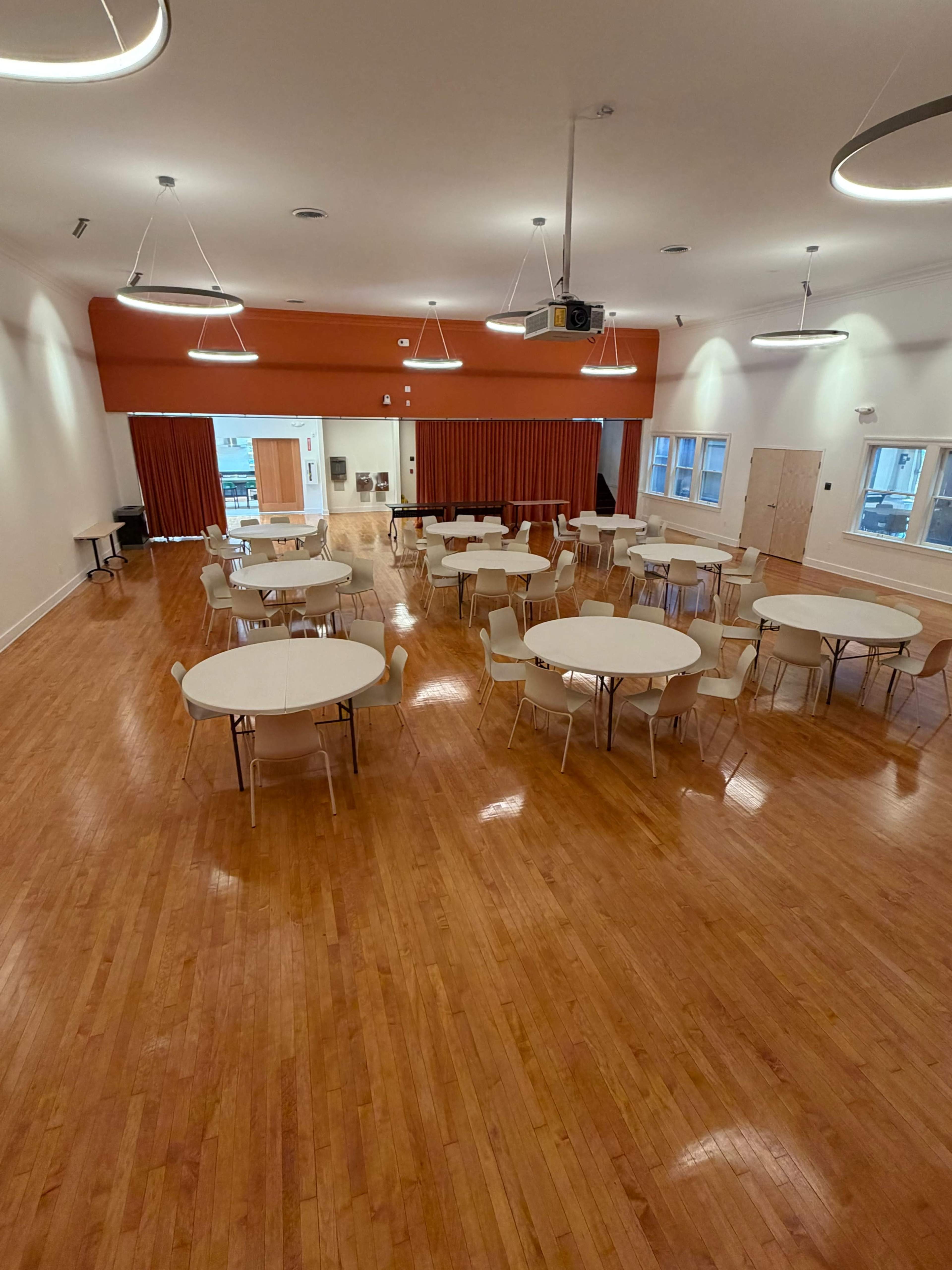 The image shows a large indoor space with several round tables and chairs arranged neatly on a polished wooden floor, with orange and white walls and modern lighting fixtures.