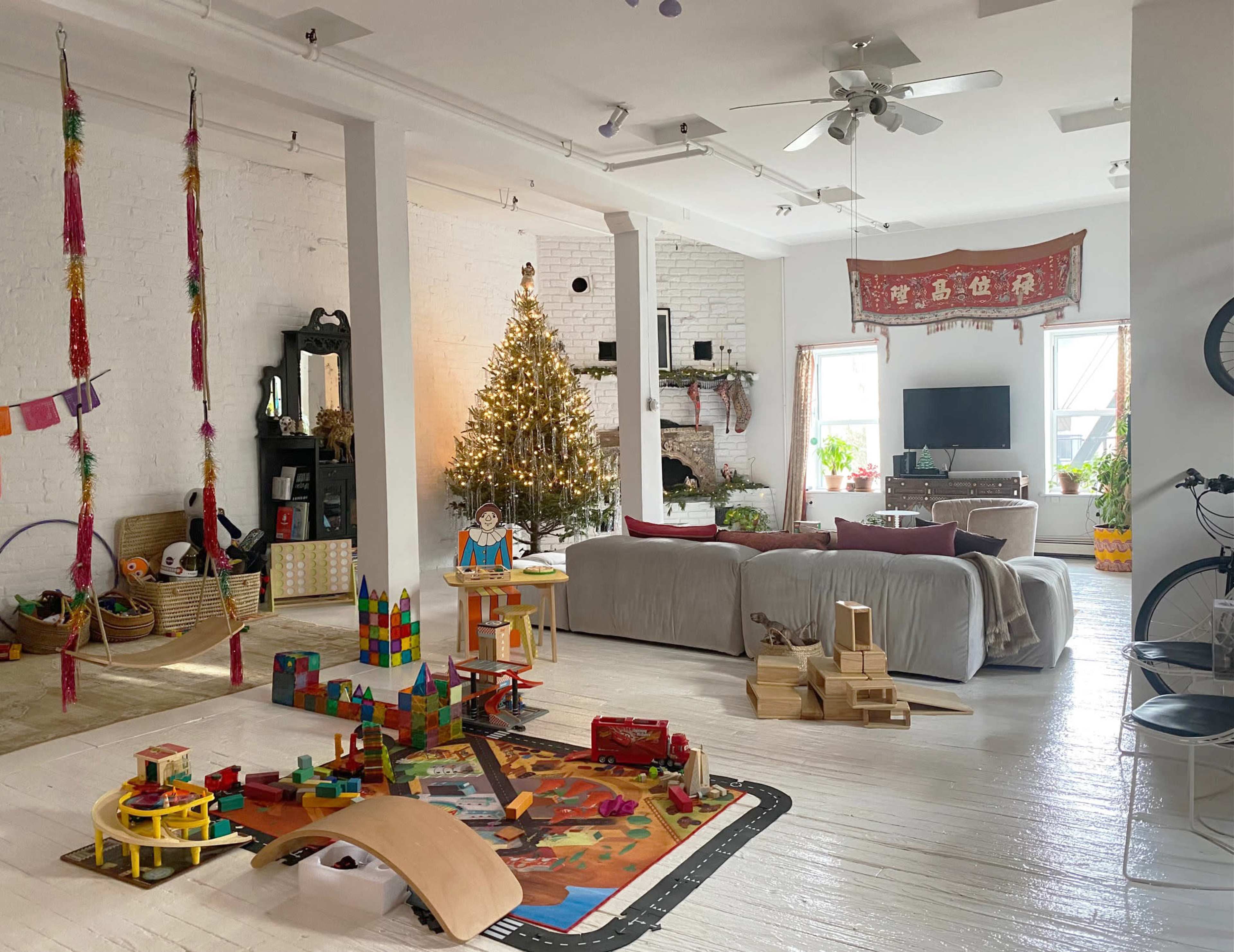 A bright living room features a Christmas tree, a play area with toys, and a cozy seating arrangement with a television in the background.
