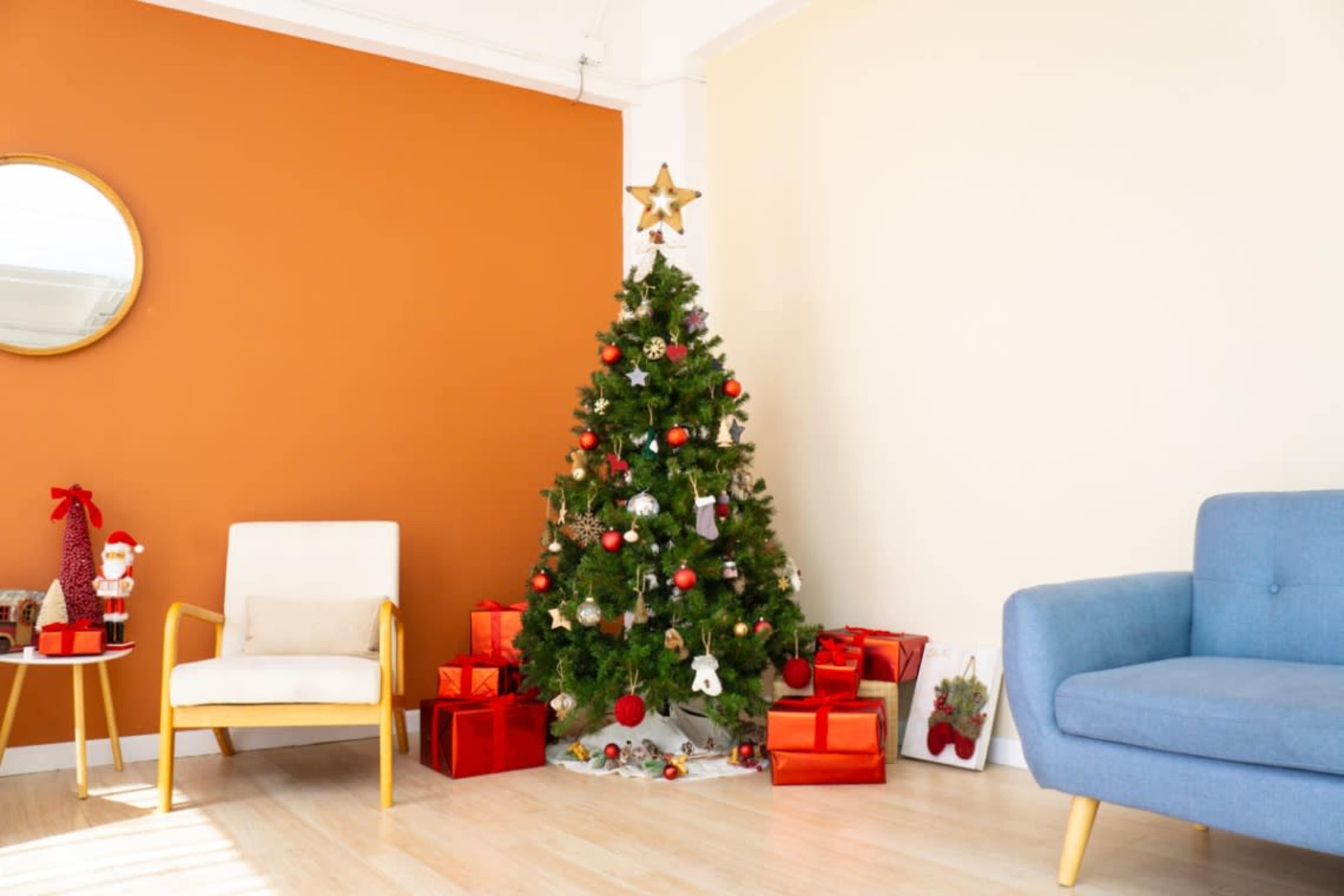 A decorated Christmas tree with presents is positioned next to a cream-colored wall in a brightly lit room.