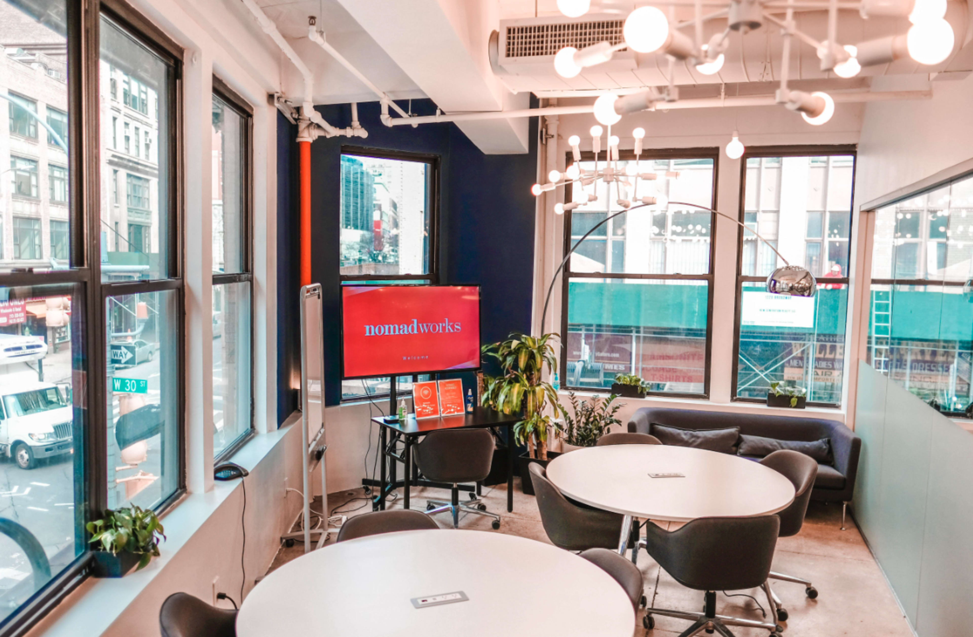 The image shows a modern coworking space with large windows, circular tables, a TV displaying "nomadworks," and a green plant in the corner.