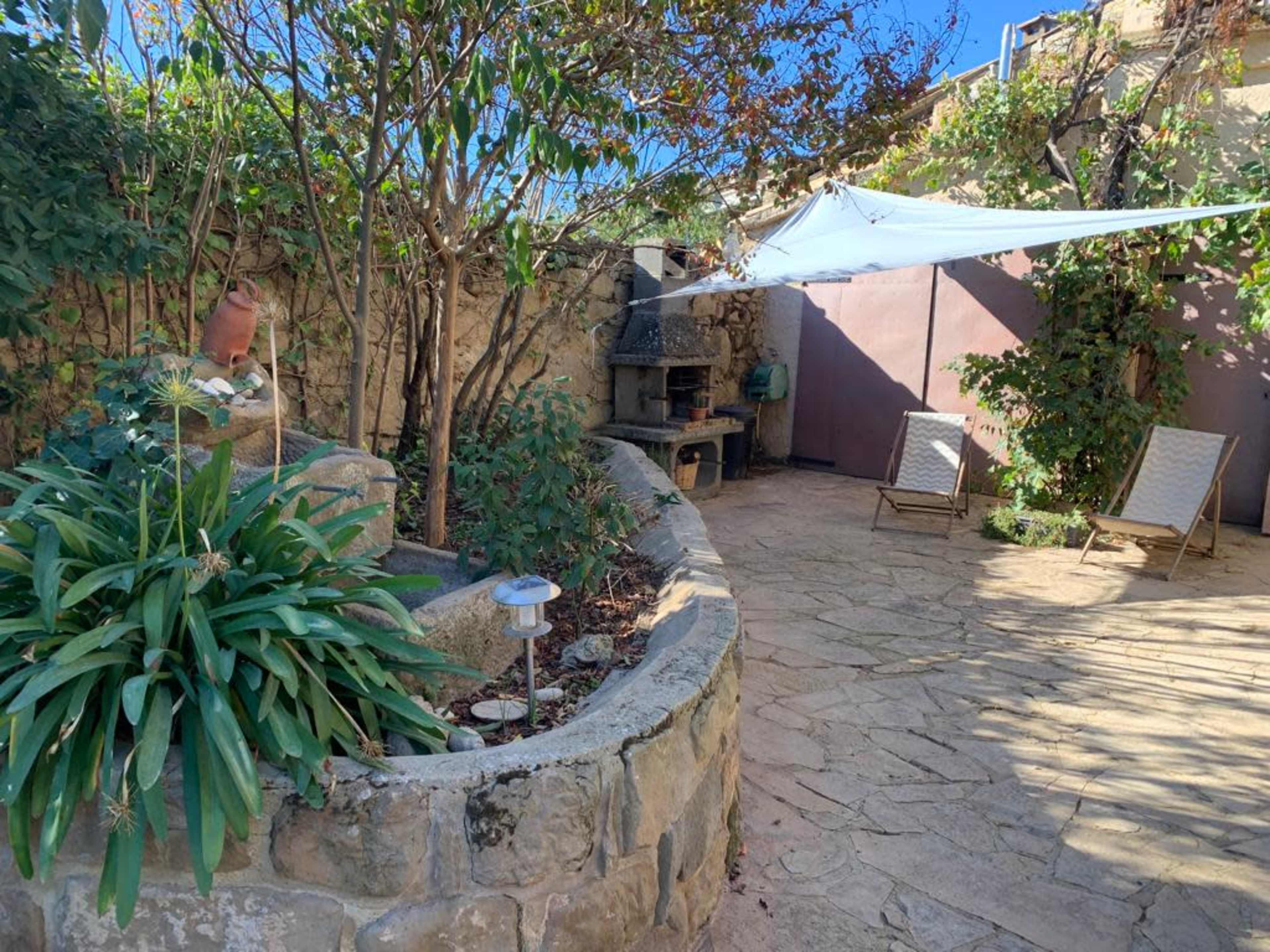 The image shows a stone-paved garden area featuring a seating space, a stone wall, and a shaded section with a white canopy.