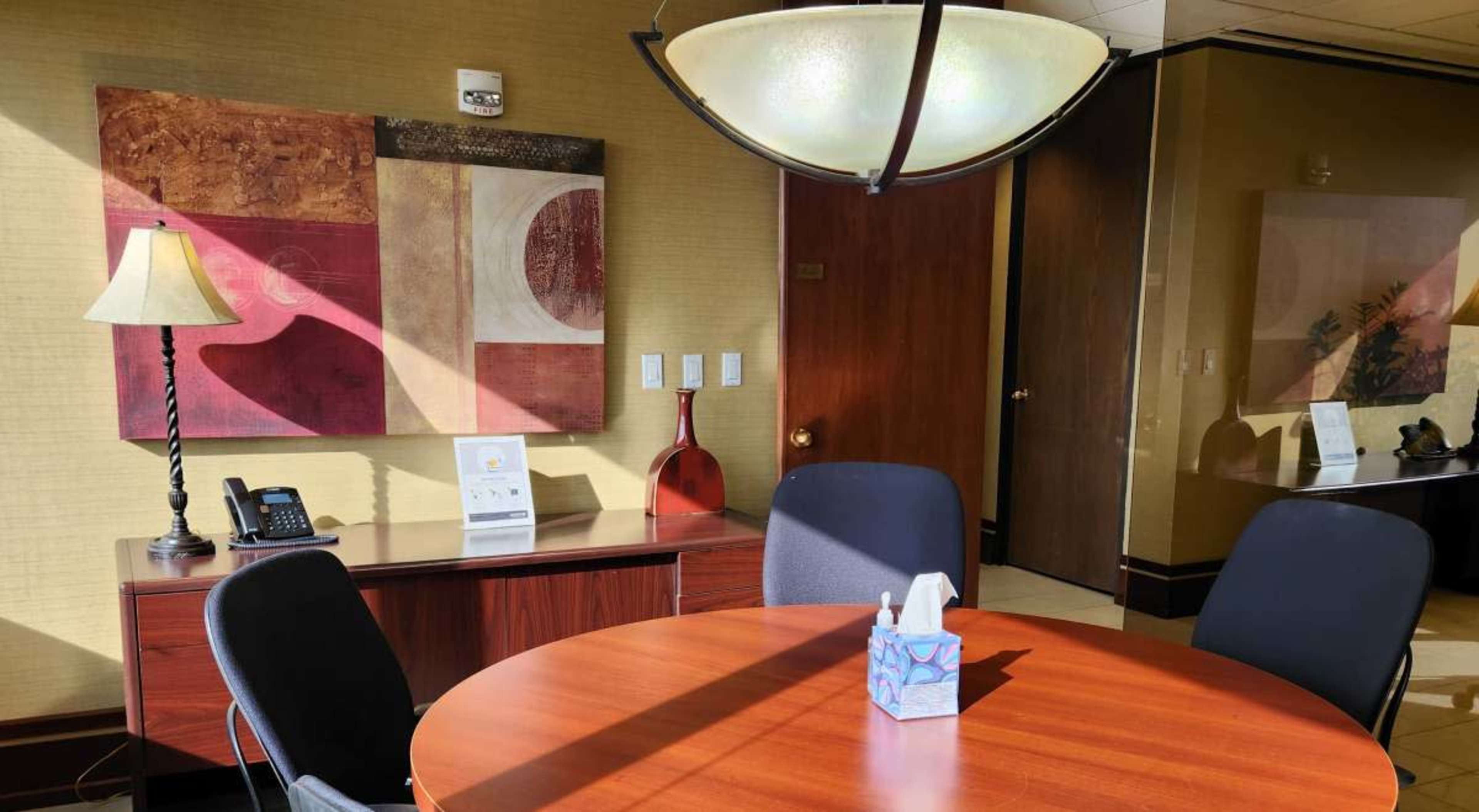 A well-lit office space featuring a round wooden table, black chairs, a lamp, and abstract wall art.