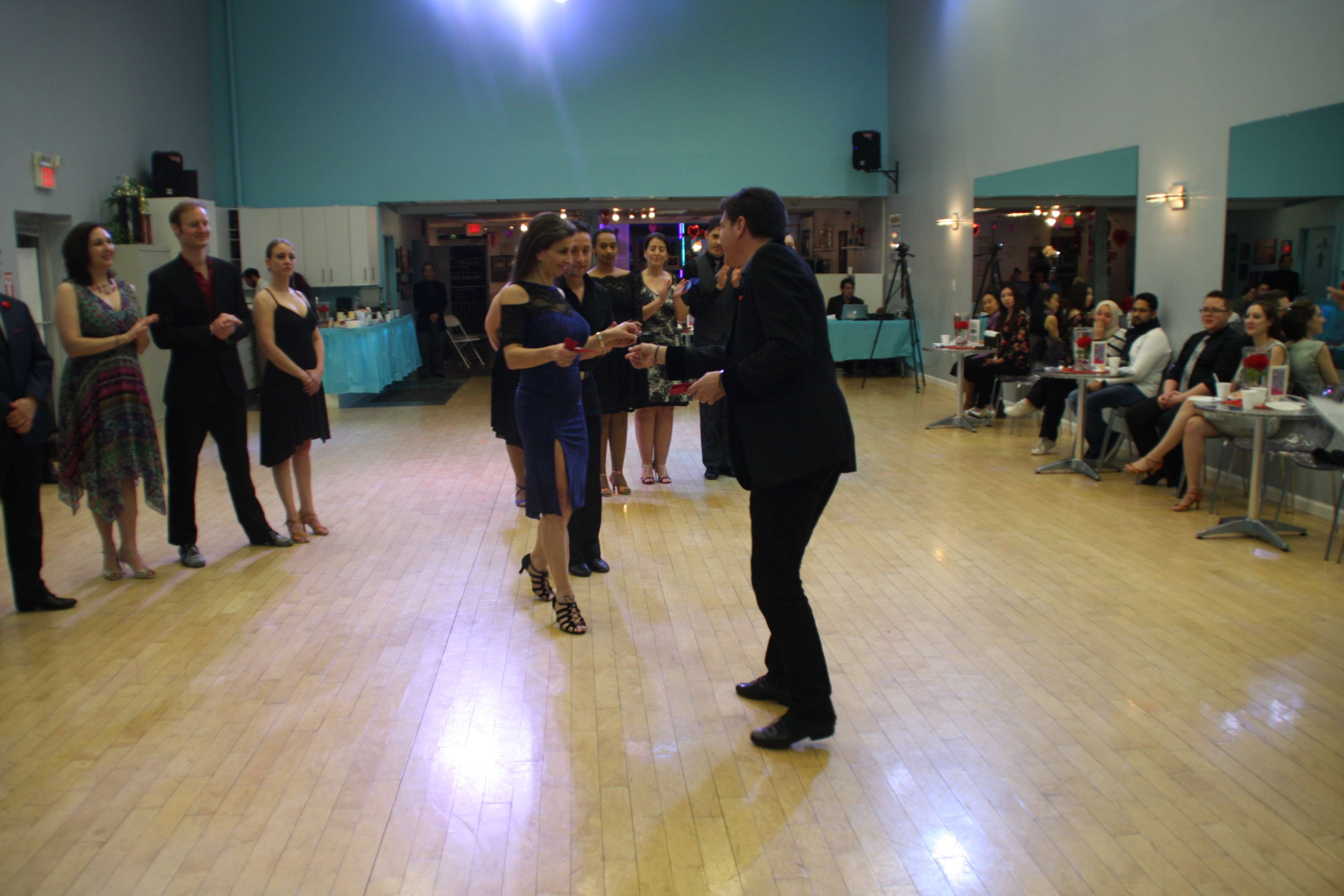 A couple dances in the center of a ballroom while spectators watch from the sides.
