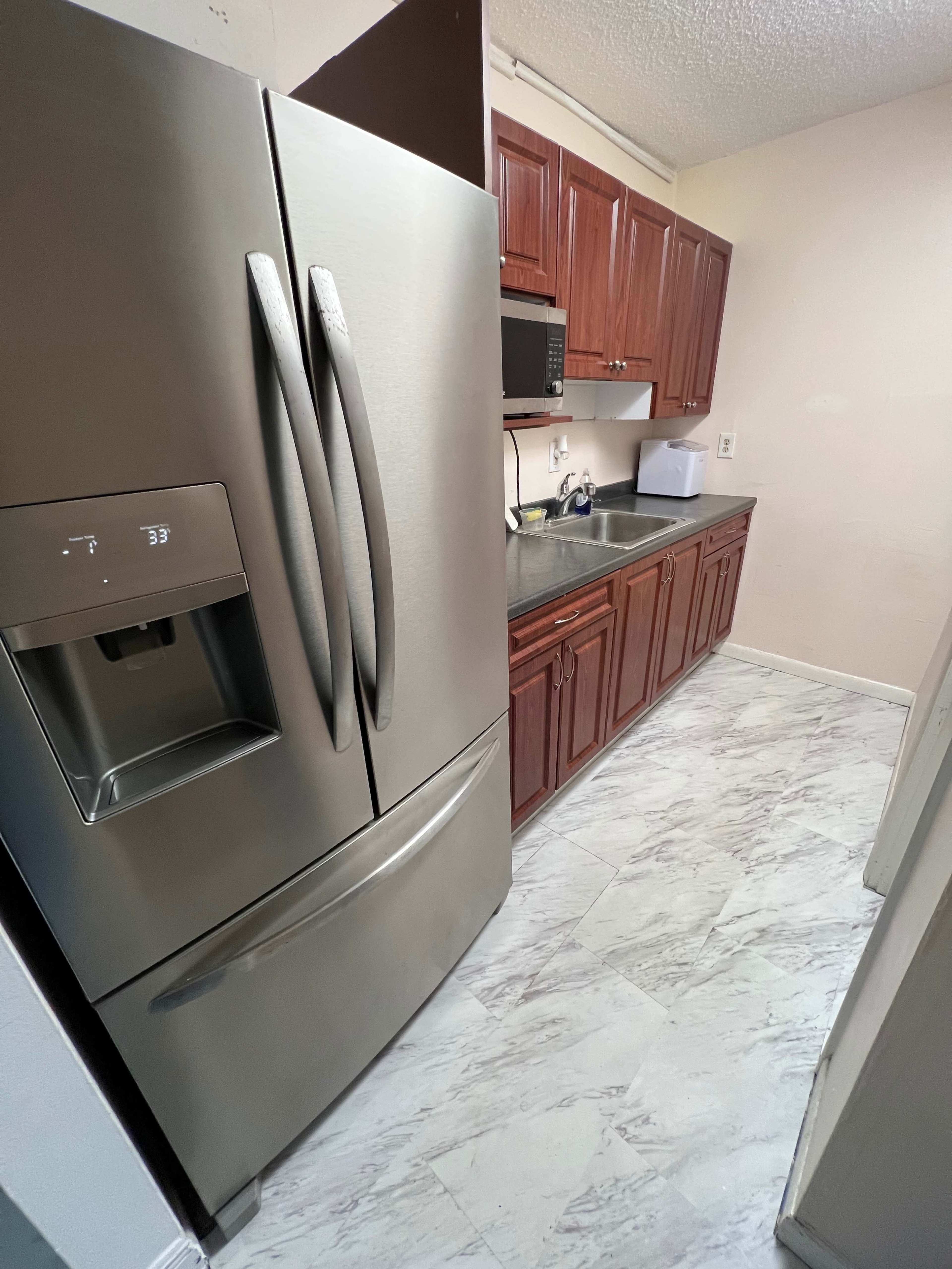 The image shows a kitchen with a silver refrigerator on the left and wooden cabinets above a countertop on the right.