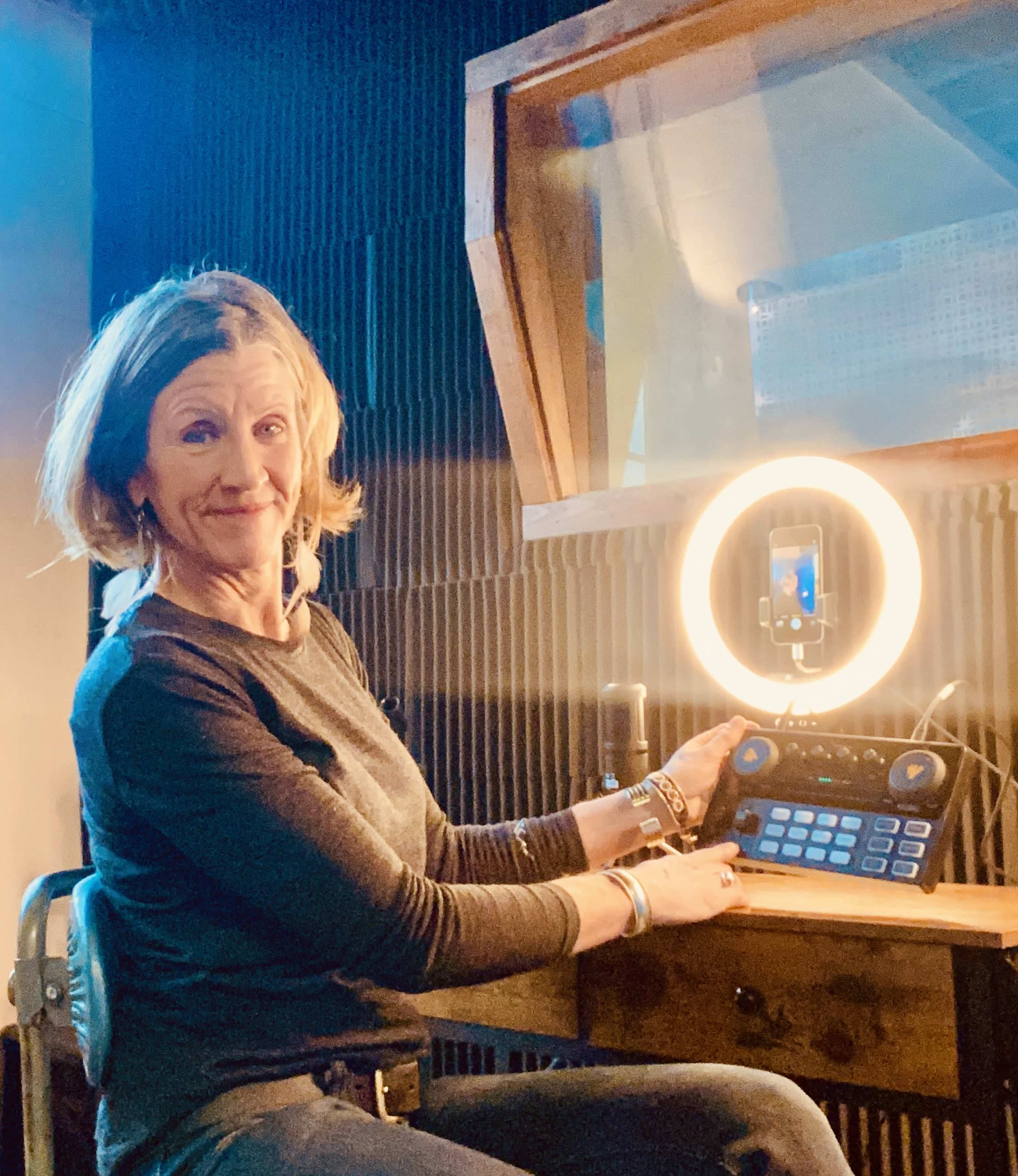 A woman sits in a recording studio, operating an audio interface while positioned in front of a ring light.