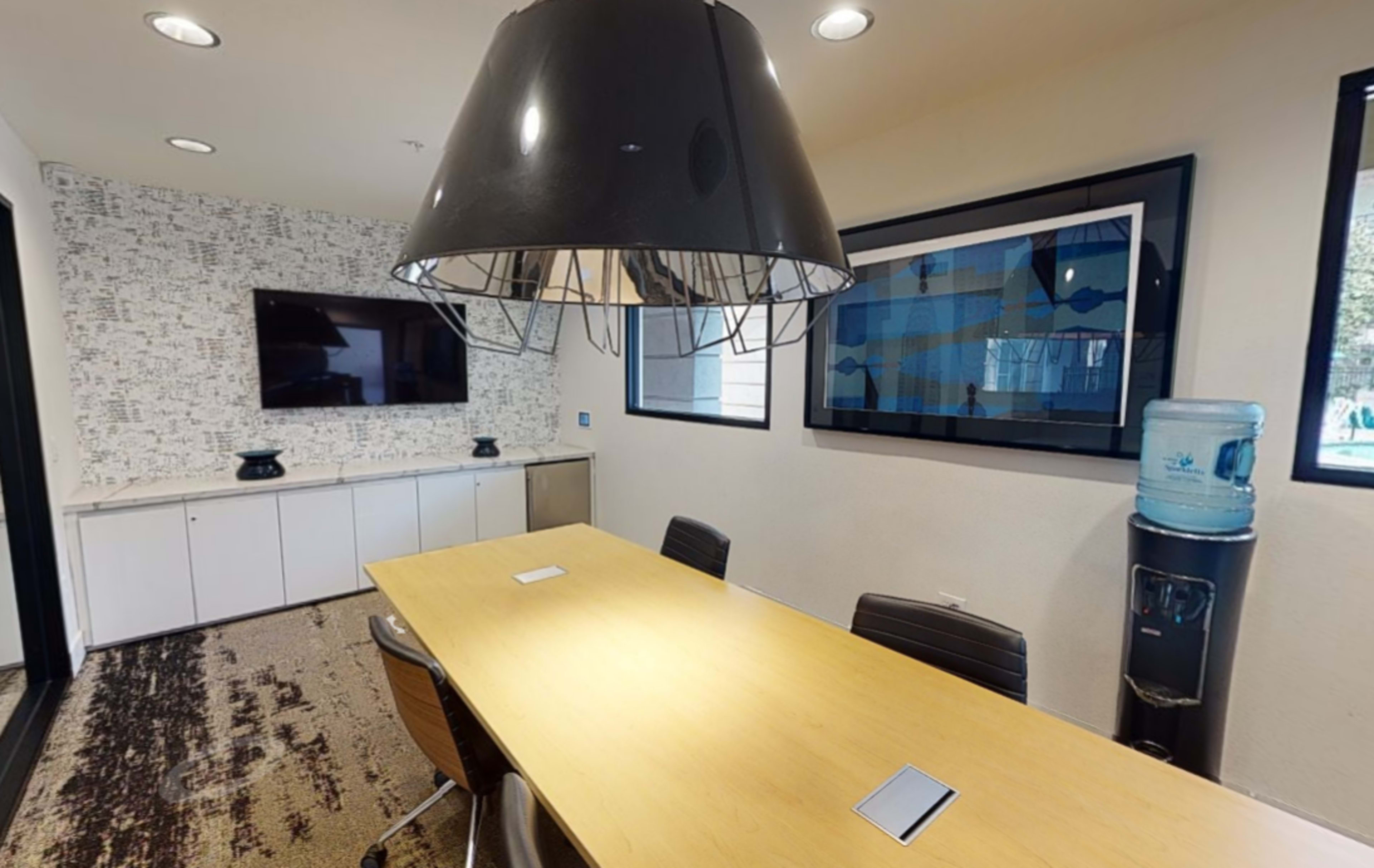 A modern conference room features a large wooden table, a water cooler, and a flat-screen television on the wall.
