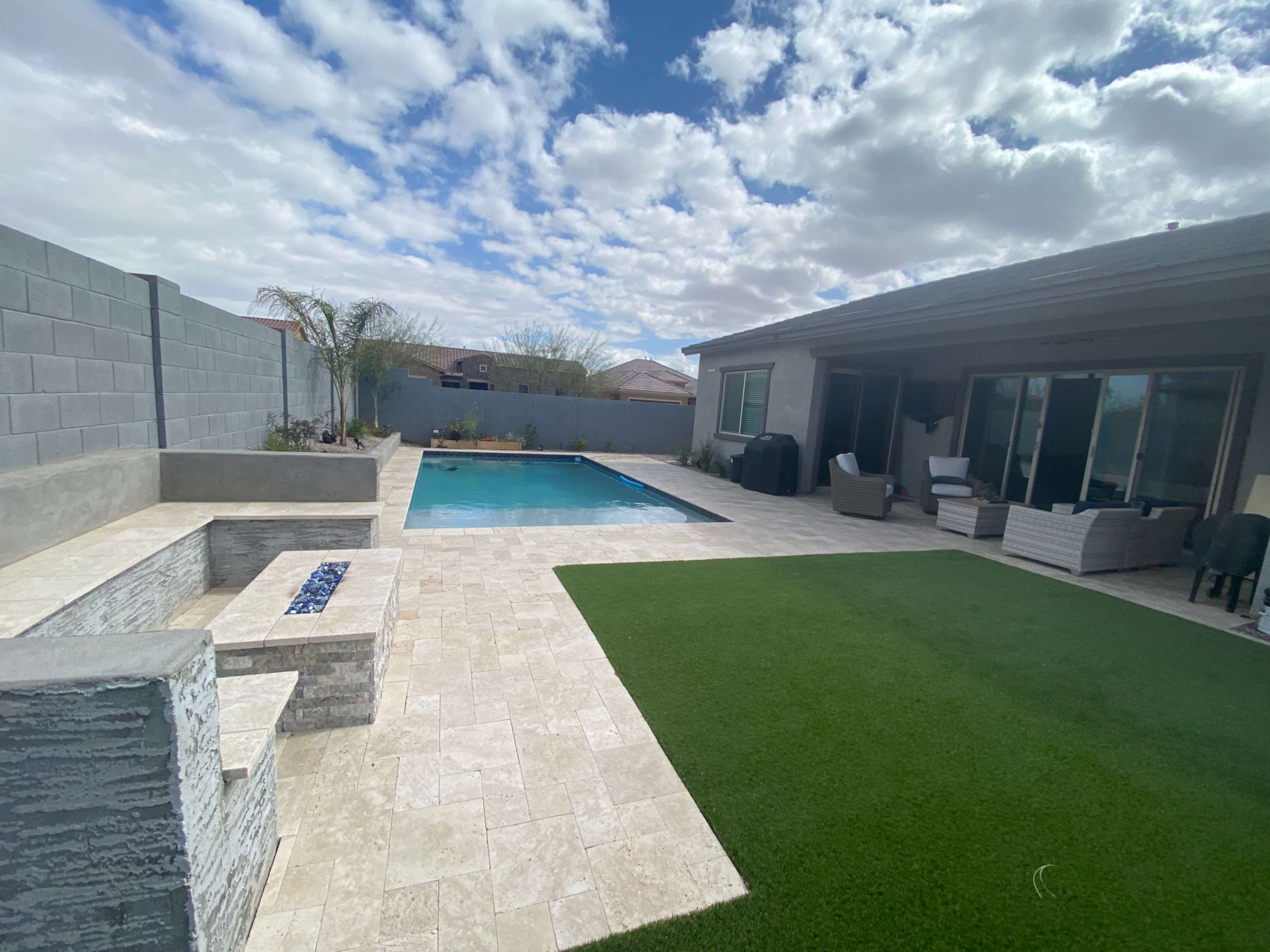 A backyard with a swimming pool, a stone fire pit, and a well-maintained lawn, surrounded by a low wall and a house.