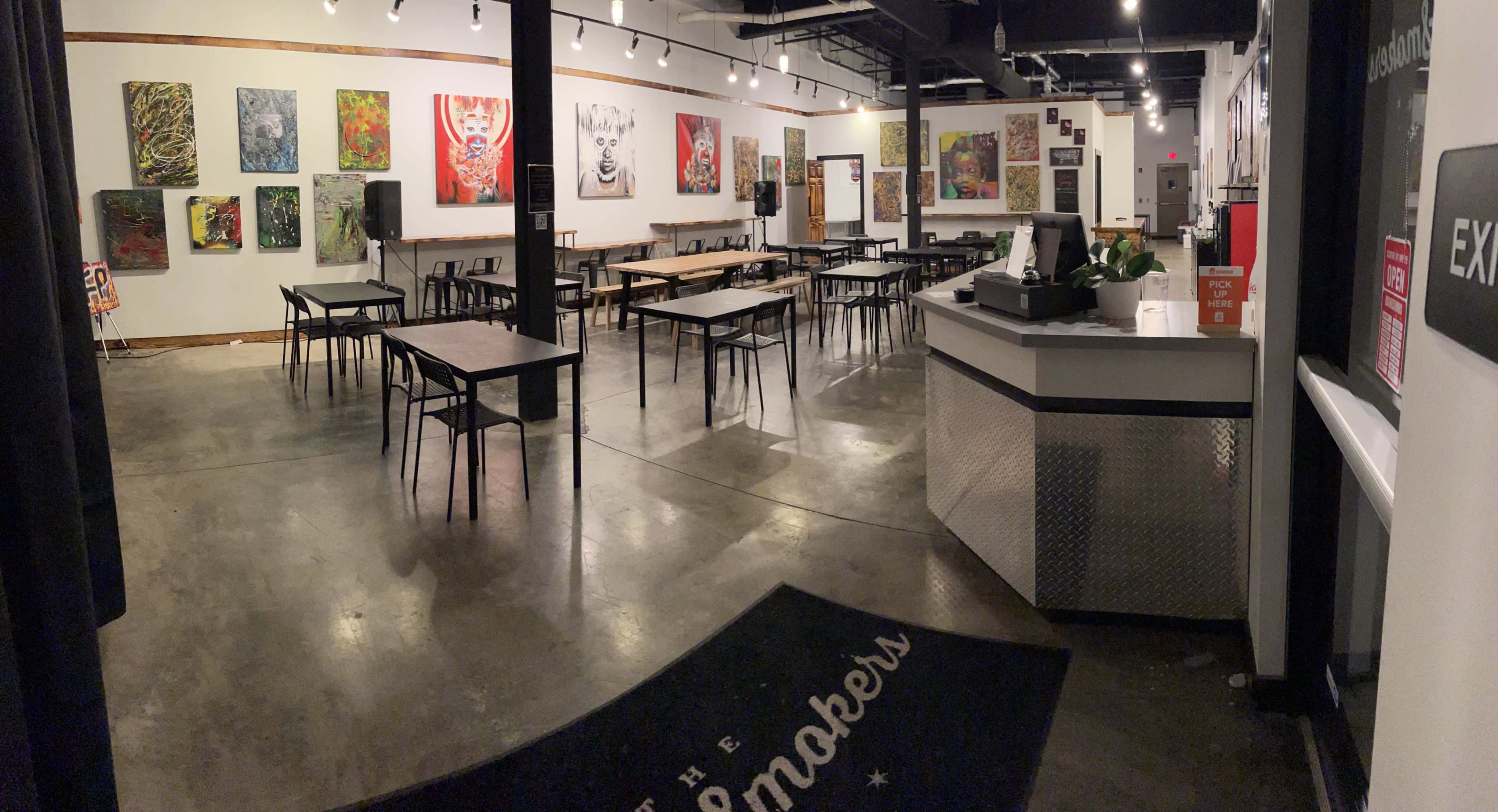A spacious art gallery with vibrant paintings on the walls, several tables arranged across the floor, and a reception area in the corner.