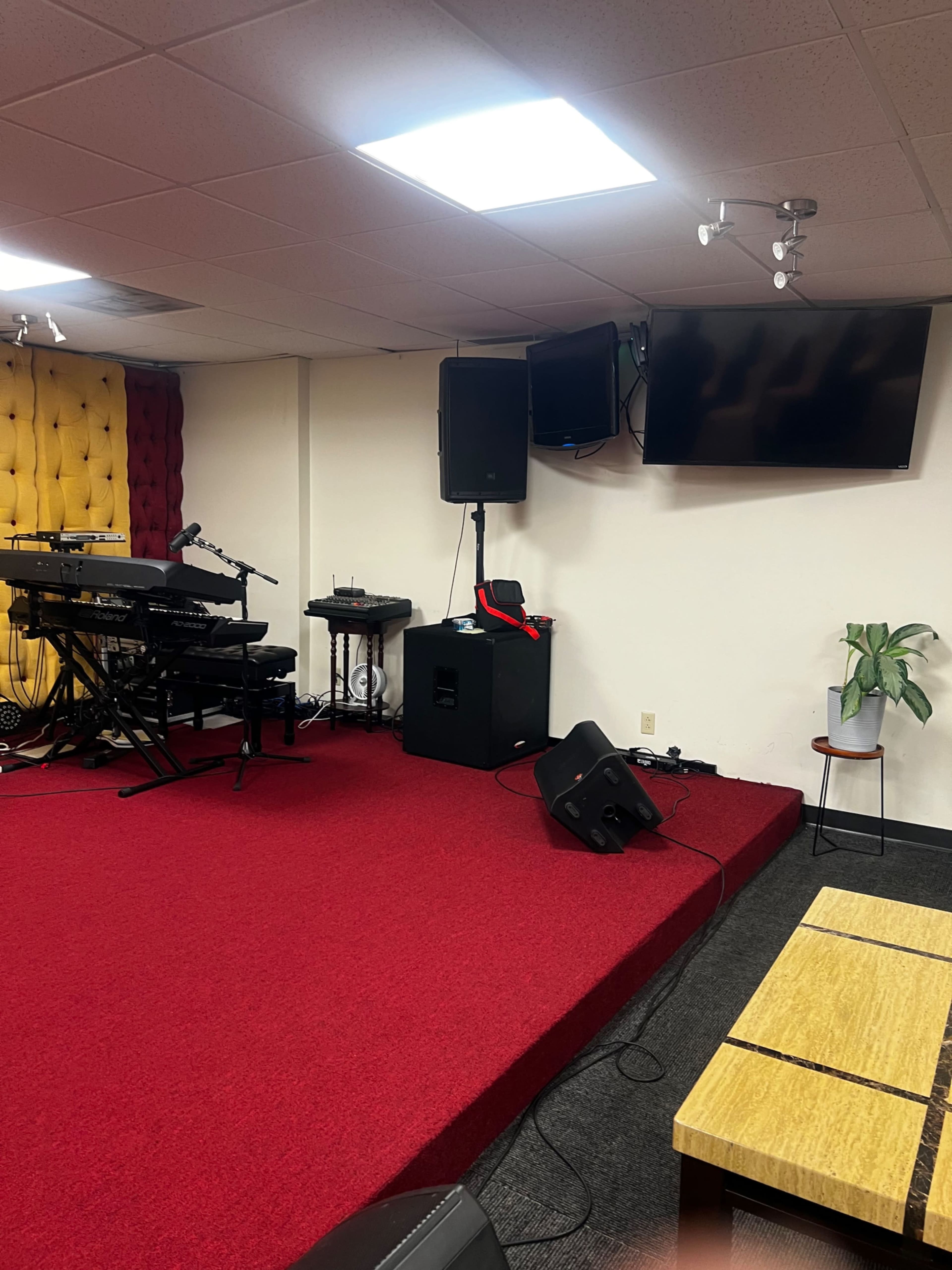 The image shows a small stage area with musical equipment, red carpet flooring, and a chair next to a potted plant in a room designed for events or performances.