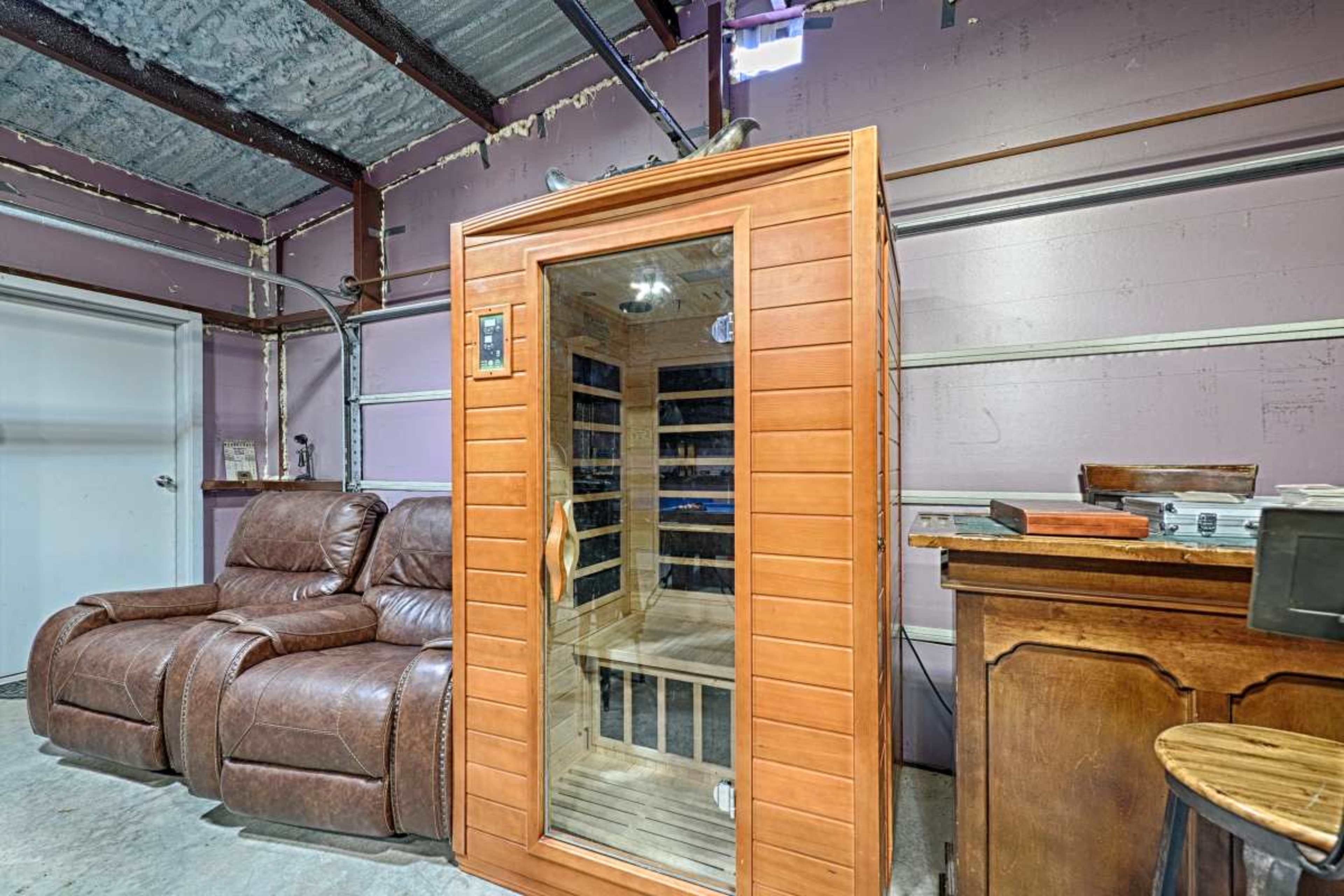 The image shows a wooden sauna situated next to two brown leather recliners in a garage-like setting.