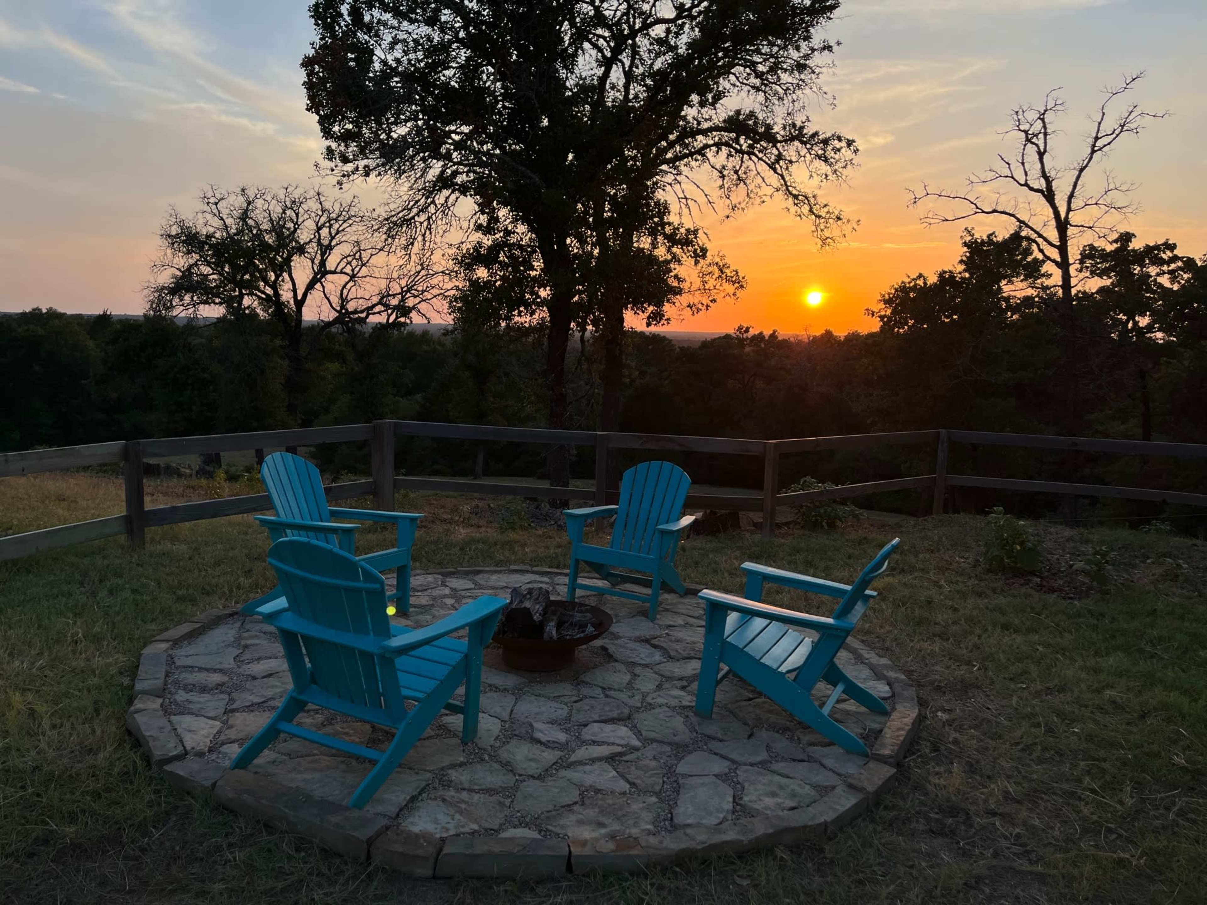 The image shows a circular fire pit area with four blue adirondack chairs, surrounded by a wooden fence, and a sunset visible in the background.