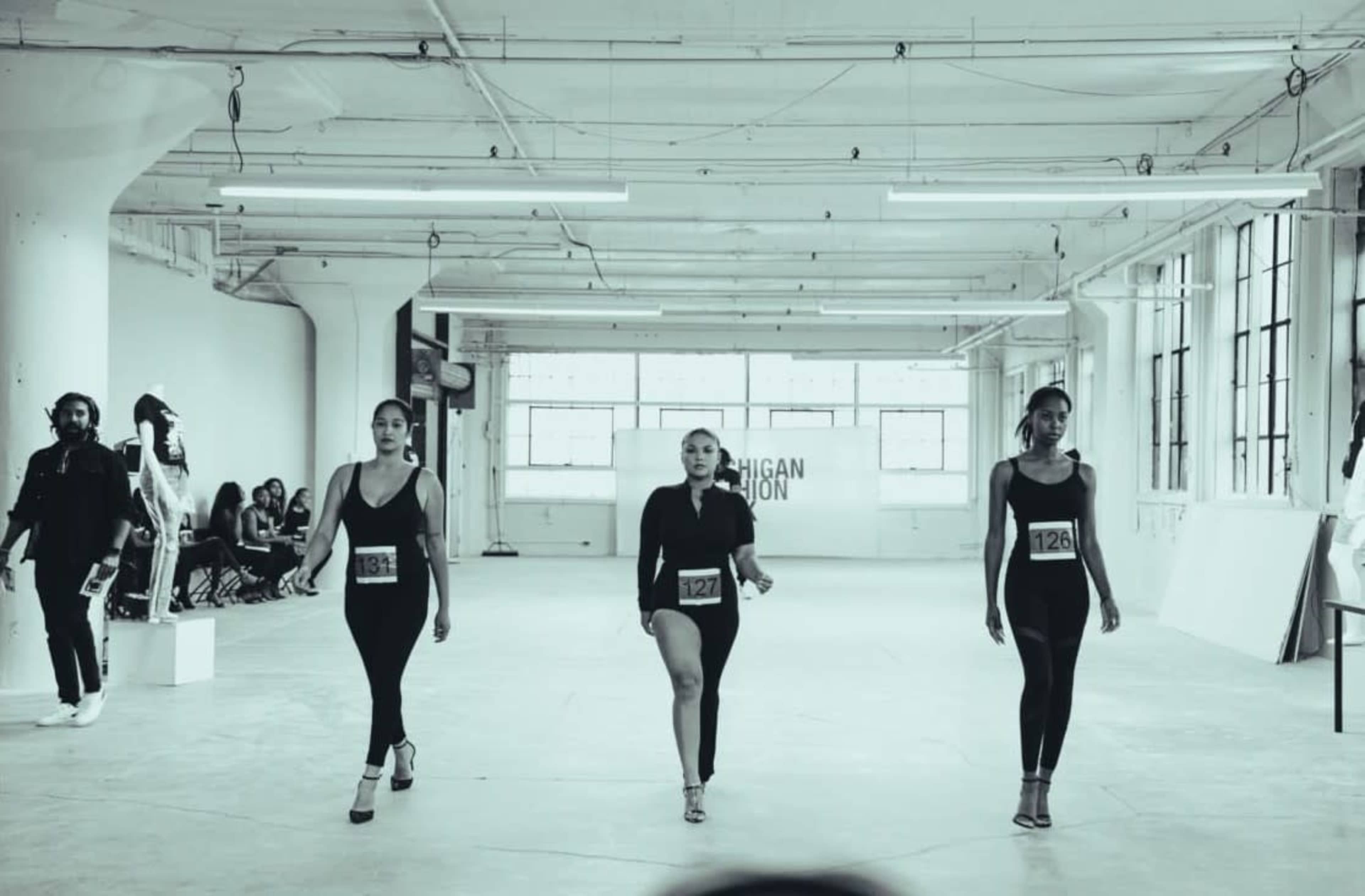 Three models walk down a spacious runway in a brightly lit studio during a fashion audition, wearing numbered tags.