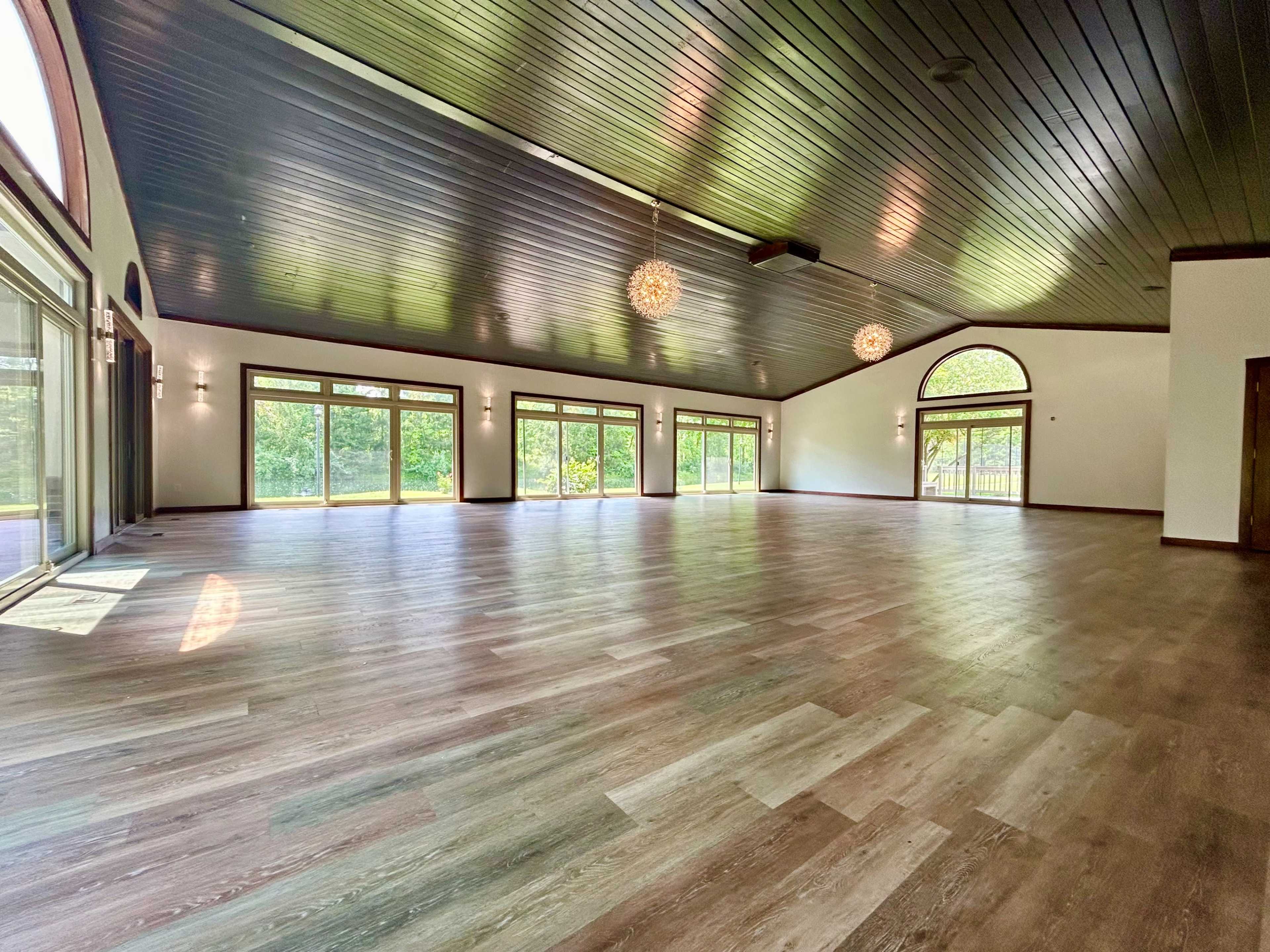 A spacious room with large windows and wooden flooring, featuring a high, dark wood ceiling and natural light flooding in from the outside.