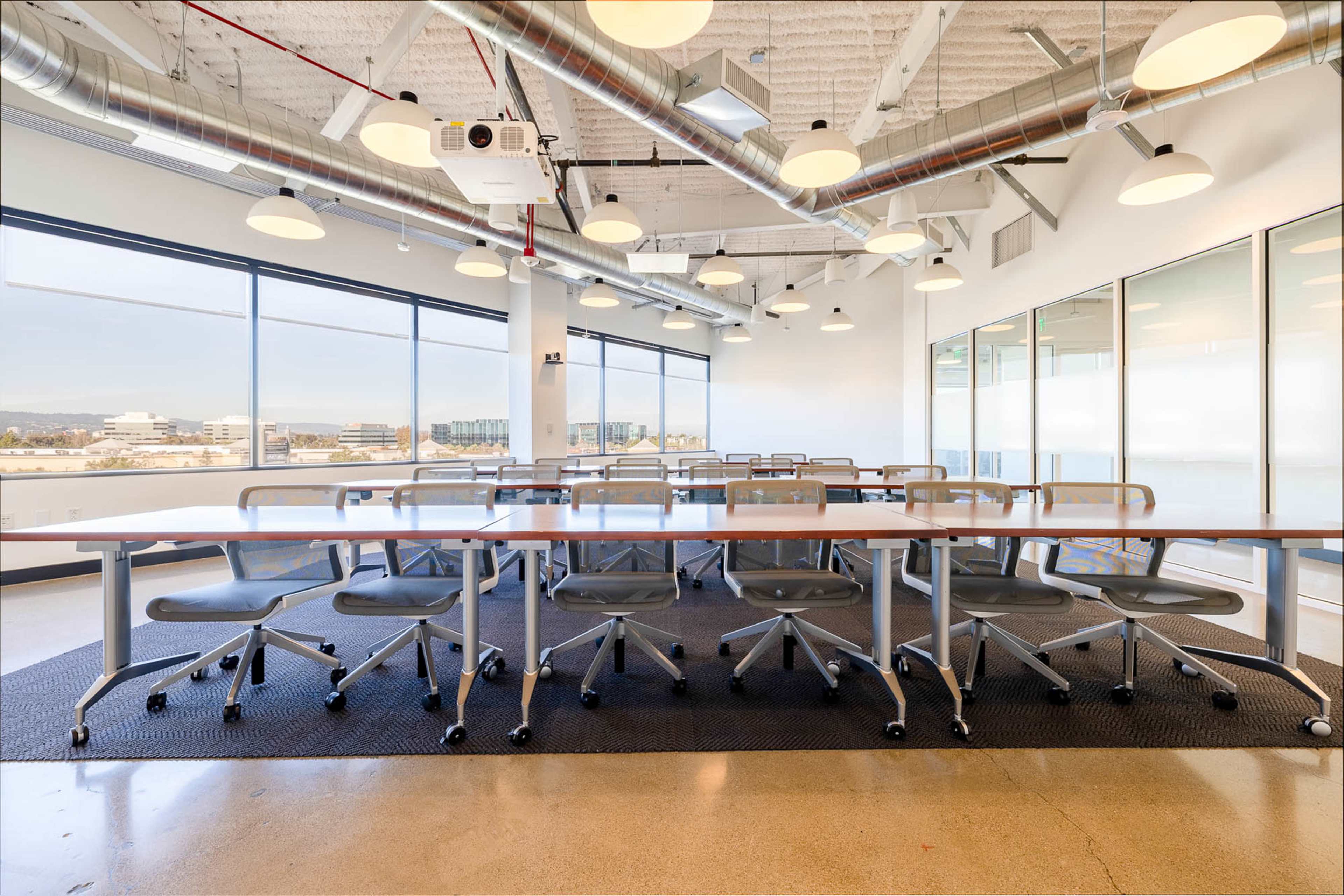 A modern conference room features a long table with rolling chairs, large windows providing natural light, and a projector mounted on the ceiling.