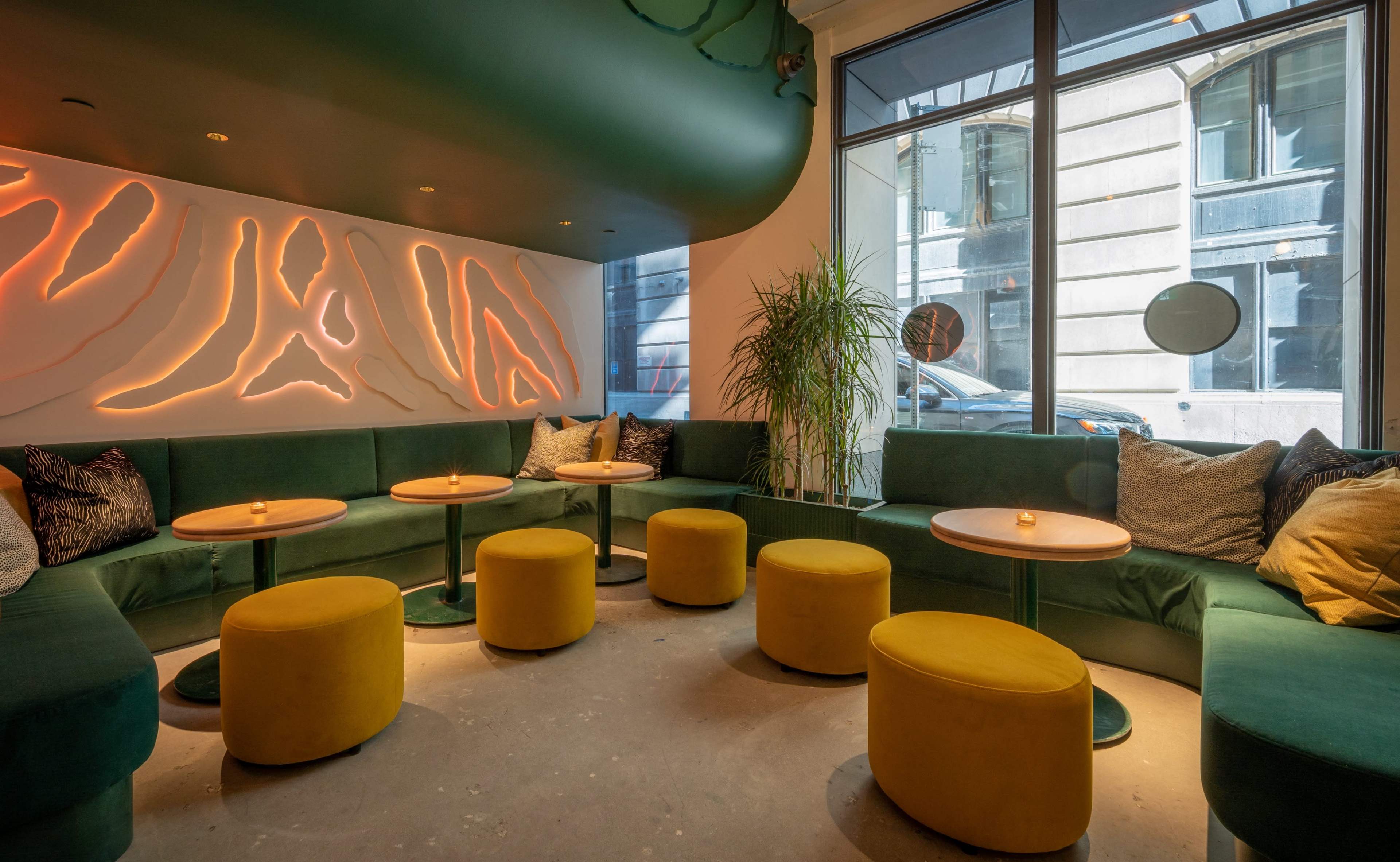 The image shows a modern lounge area with green sofas, yellow ottomans, and ambient lighting, featuring large windows that overlook a street.