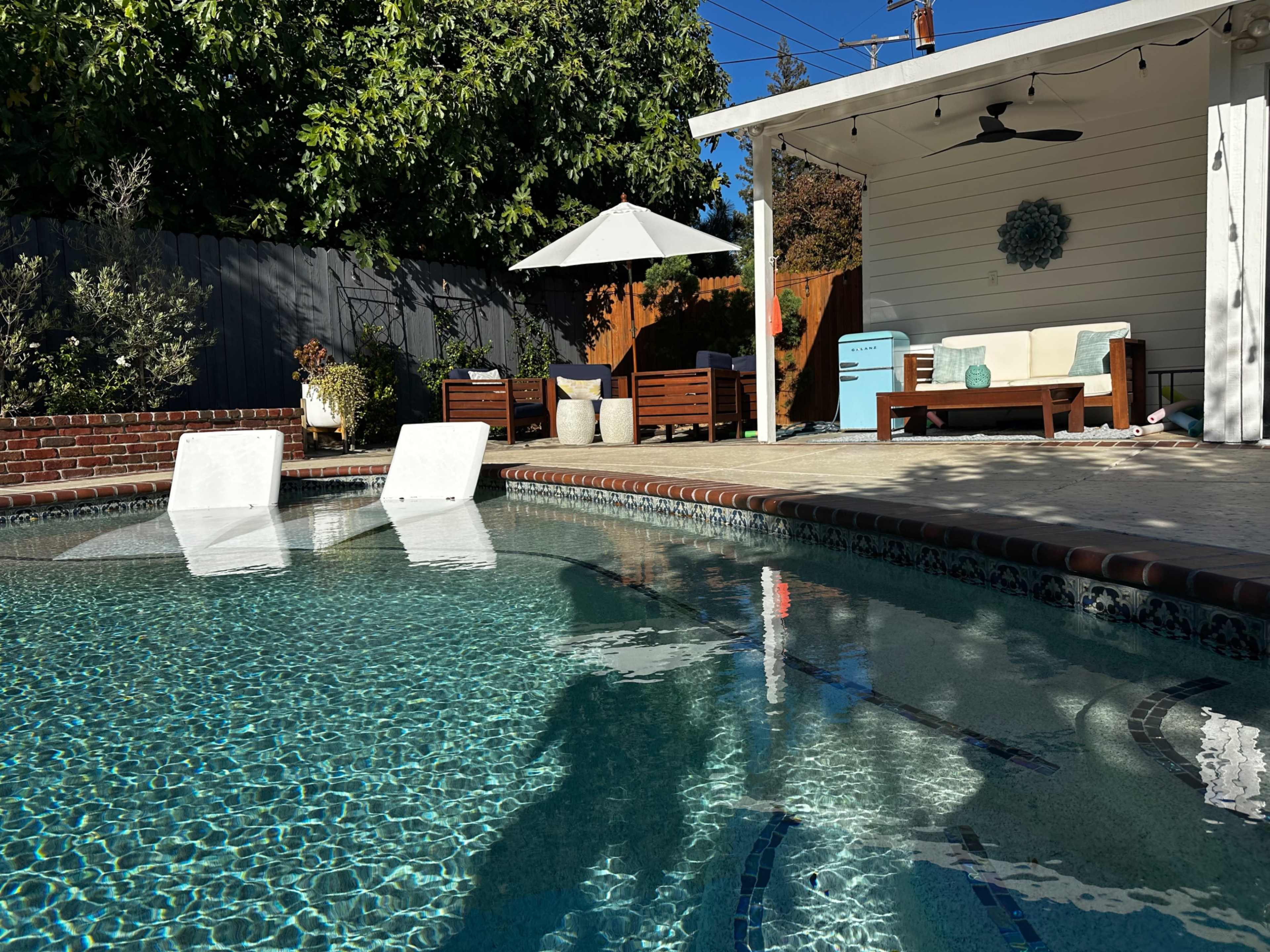 A clear swimming pool reflects the surrounding patio area, which features lounge chairs, a table with an umbrella, and outdoor furniture under a shaded porch.