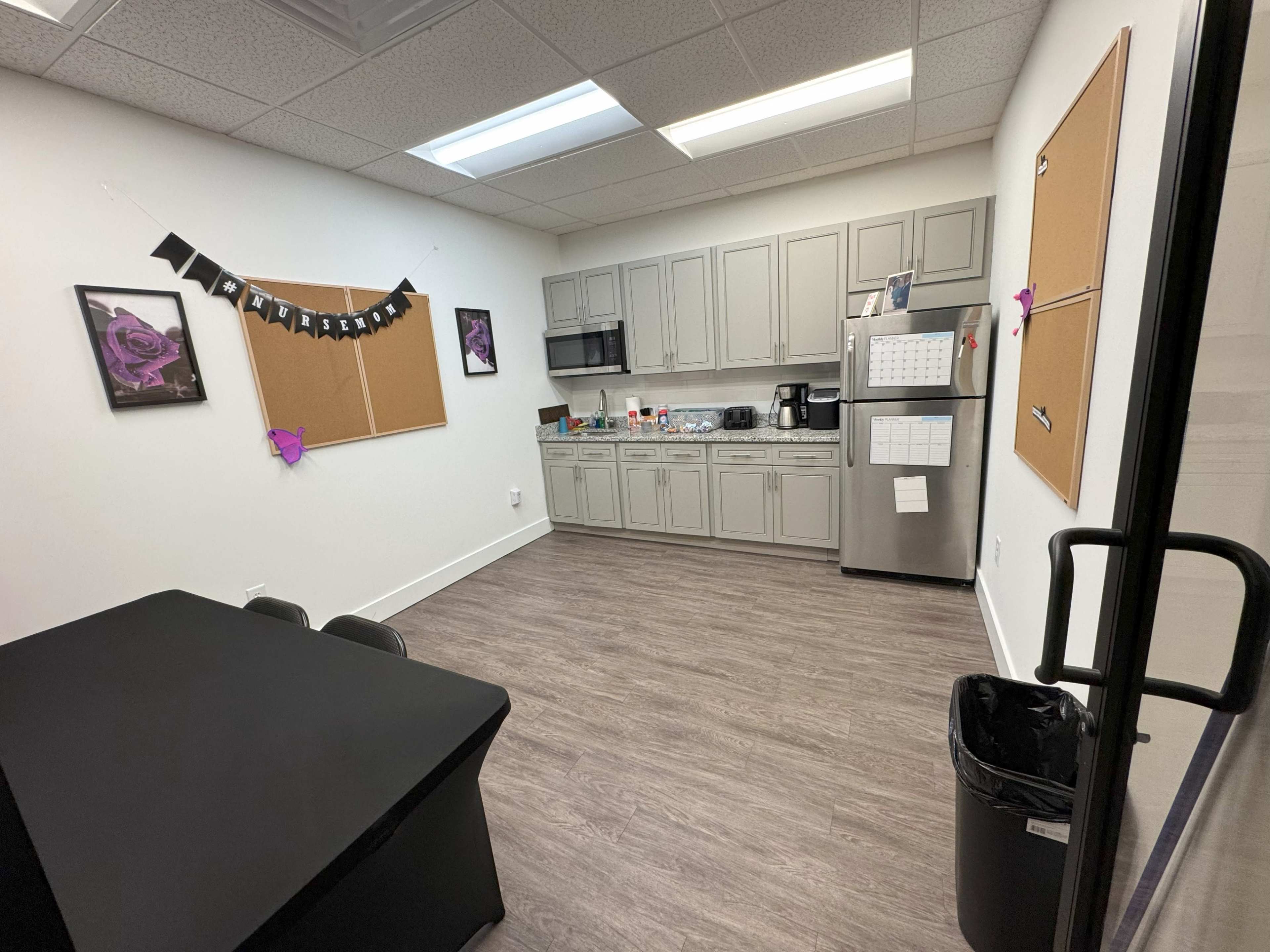 The image shows a small kitchen area with gray cabinets, a fridge, a table, and bulletin boards on the walls.