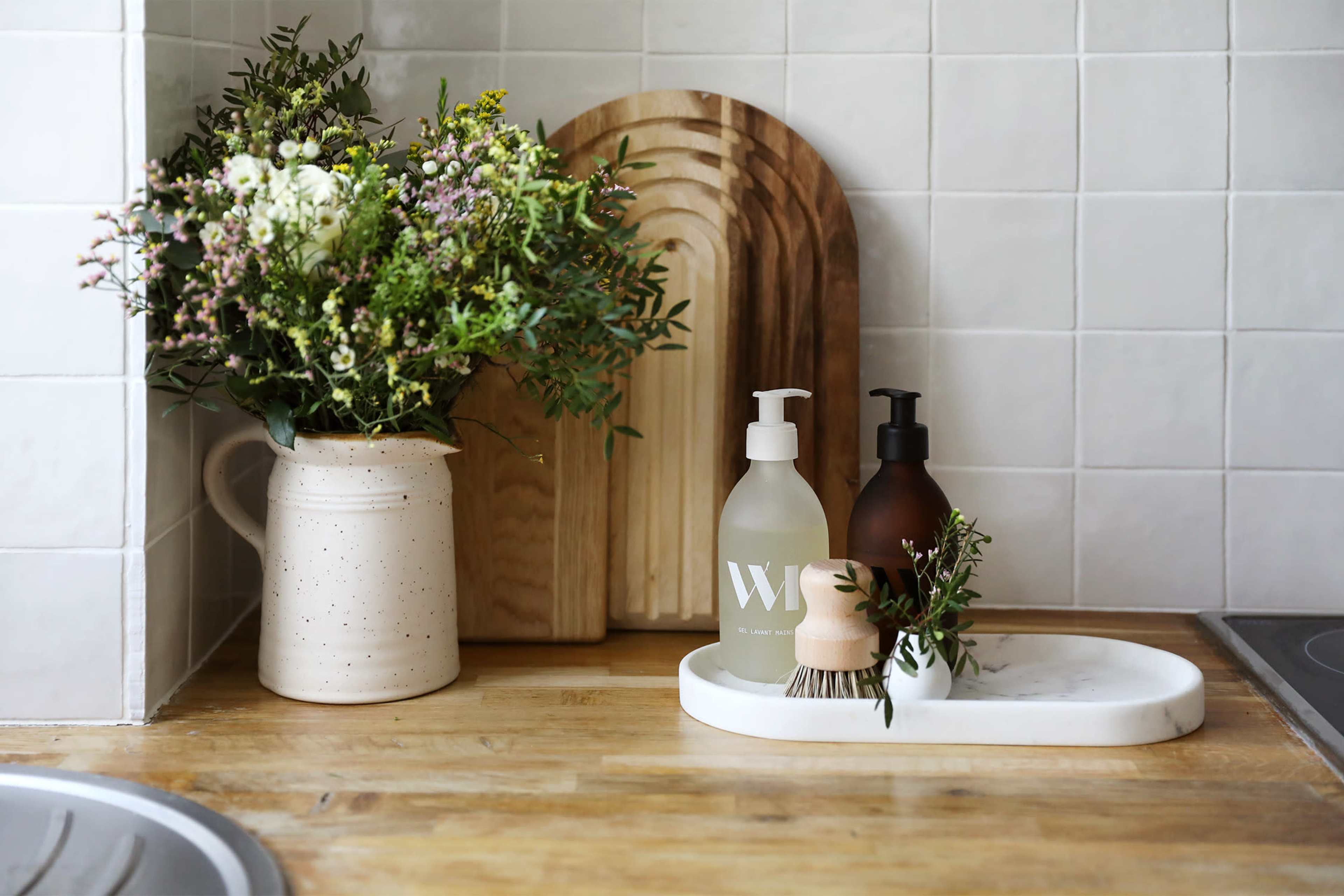 A wooden countertop displays a vase of flowers, two bottles of soap, and a small brush on a marble tray.