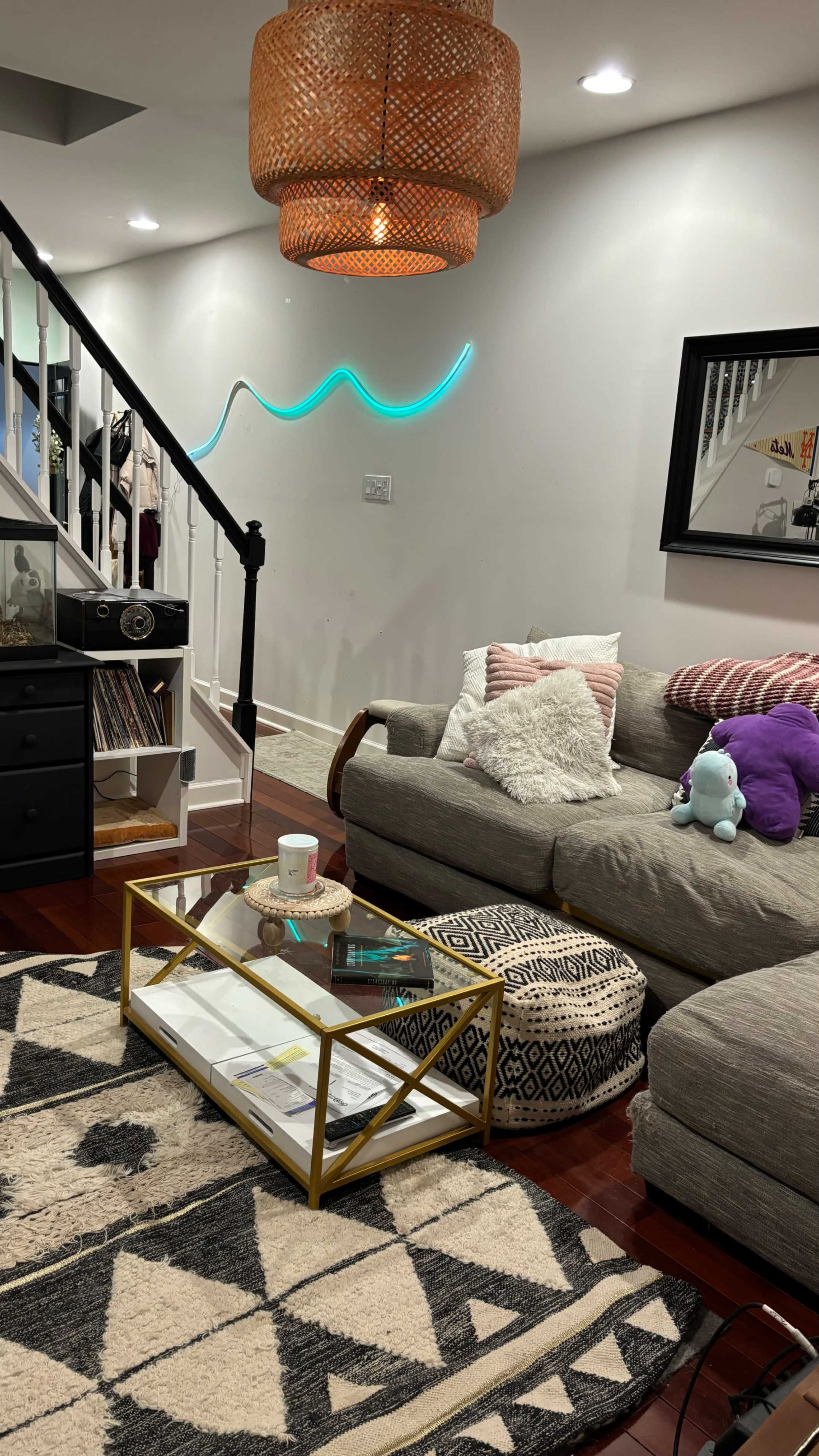 The living room features a gray couch with patterned cushions, a glass coffee table, and a staircase leading to an upper level, along with decorative elements including a pendant light and a neon wall fixture.