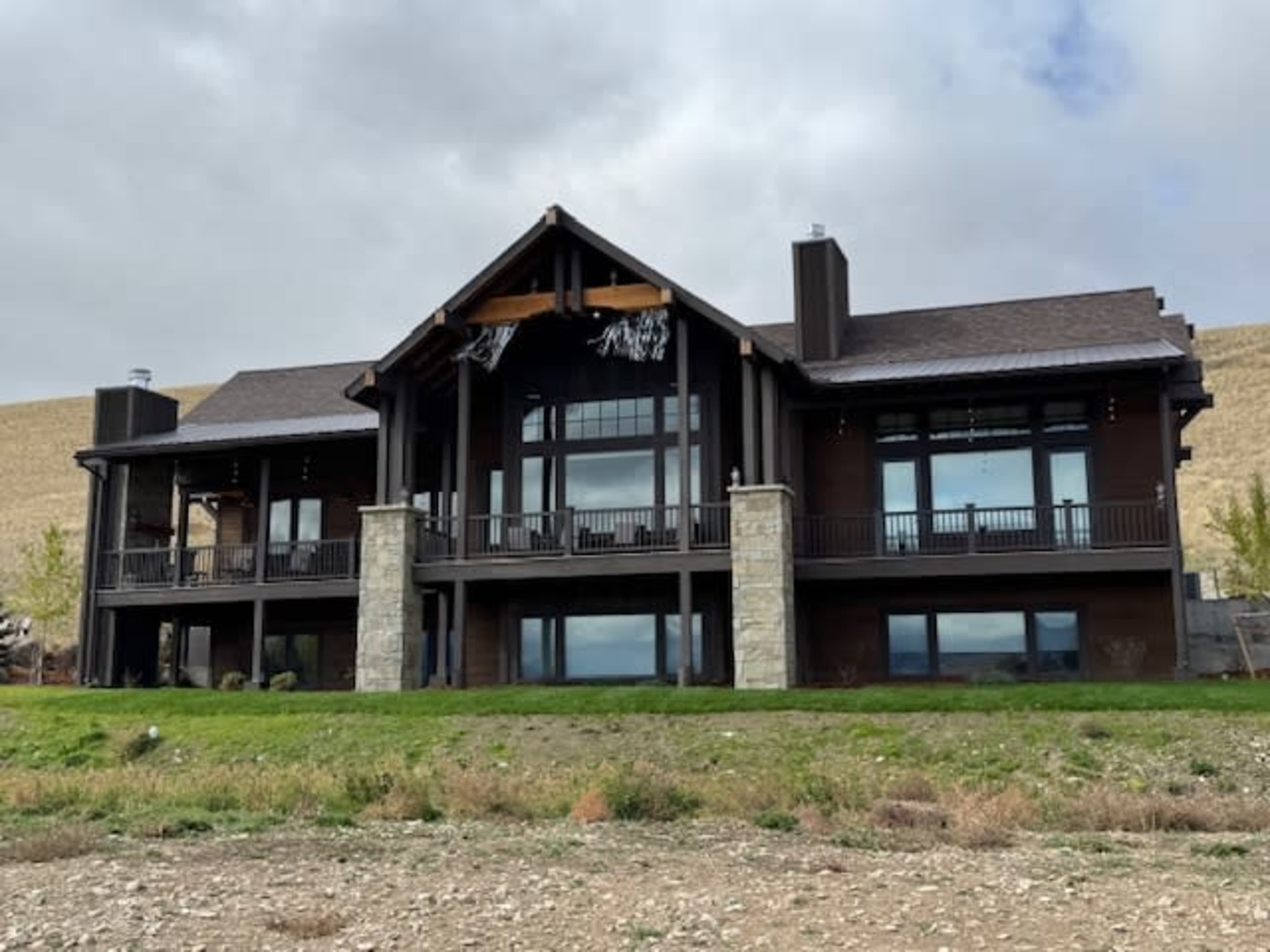 The image shows a large, two-story house with a steeply pitched roof, large windows, and stone accents, set against a backdrop of rolling hills under a cloudy sky.