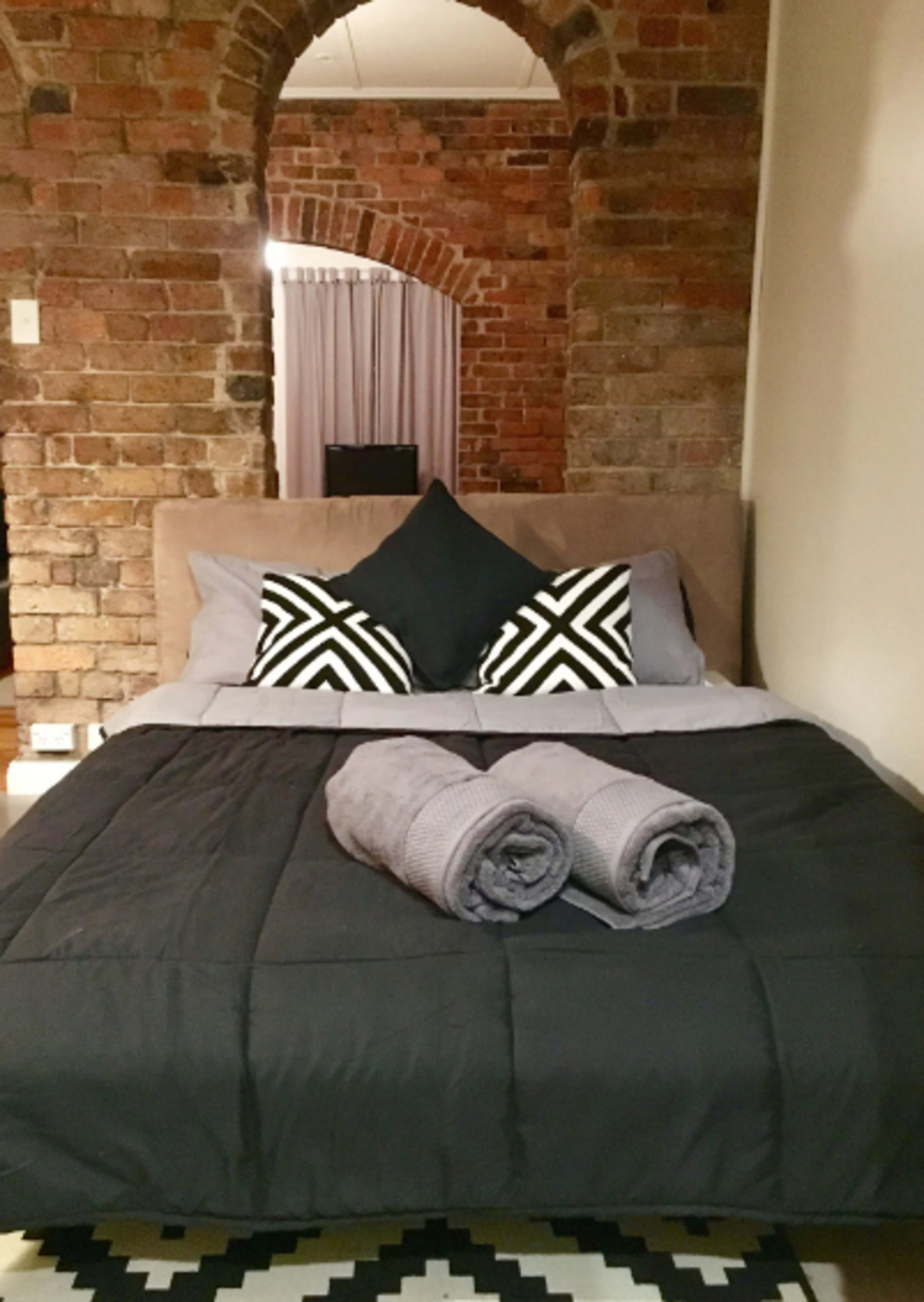 A neatly made bed with black and gray linens is positioned against a brick wall archway, featuring decorative pillows and rolled towels on top.