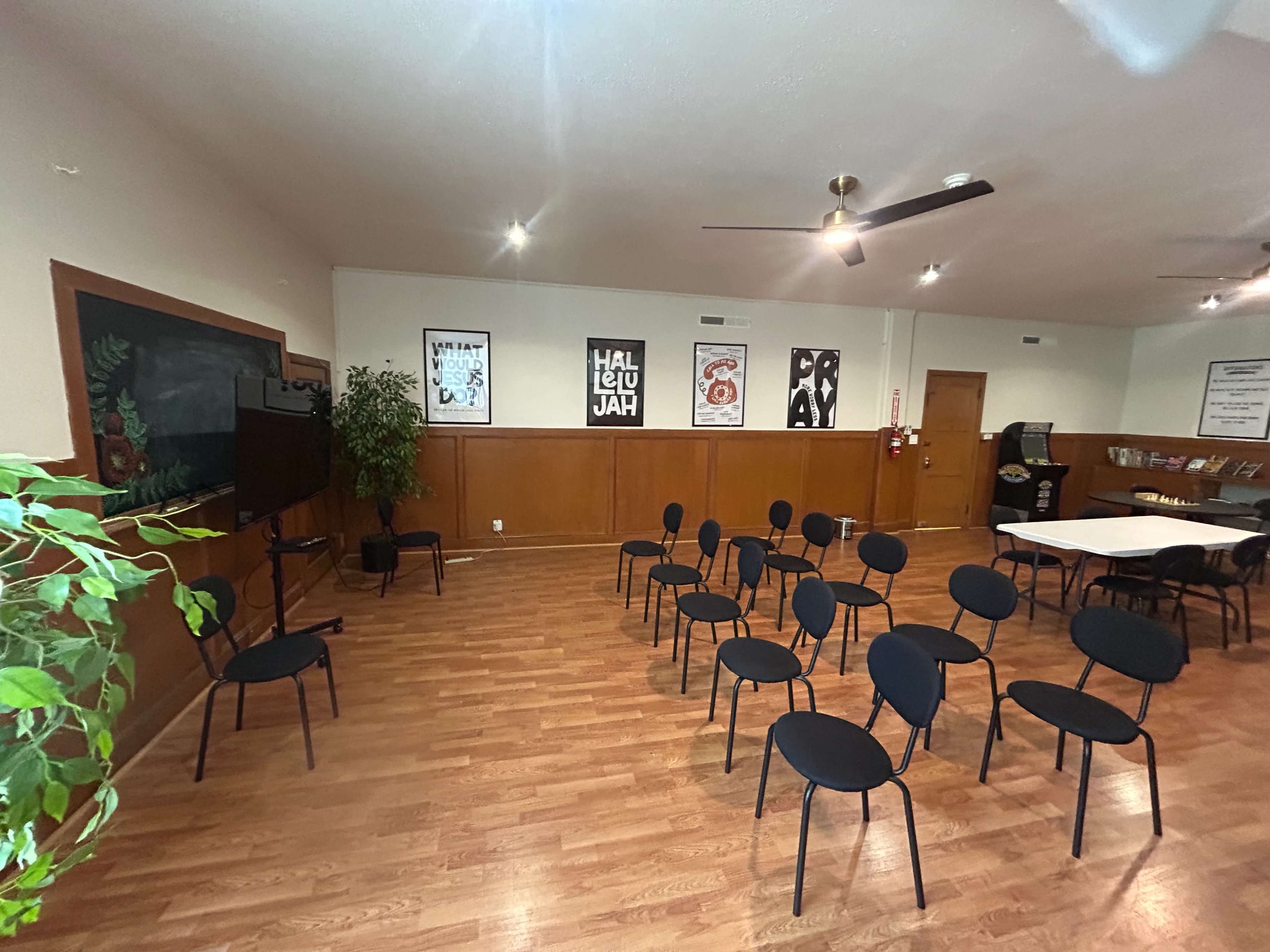 The image shows a meeting room with black chairs arranged in rows, a television on one wall, and posters hanging on another, all situated on a wooden floor.