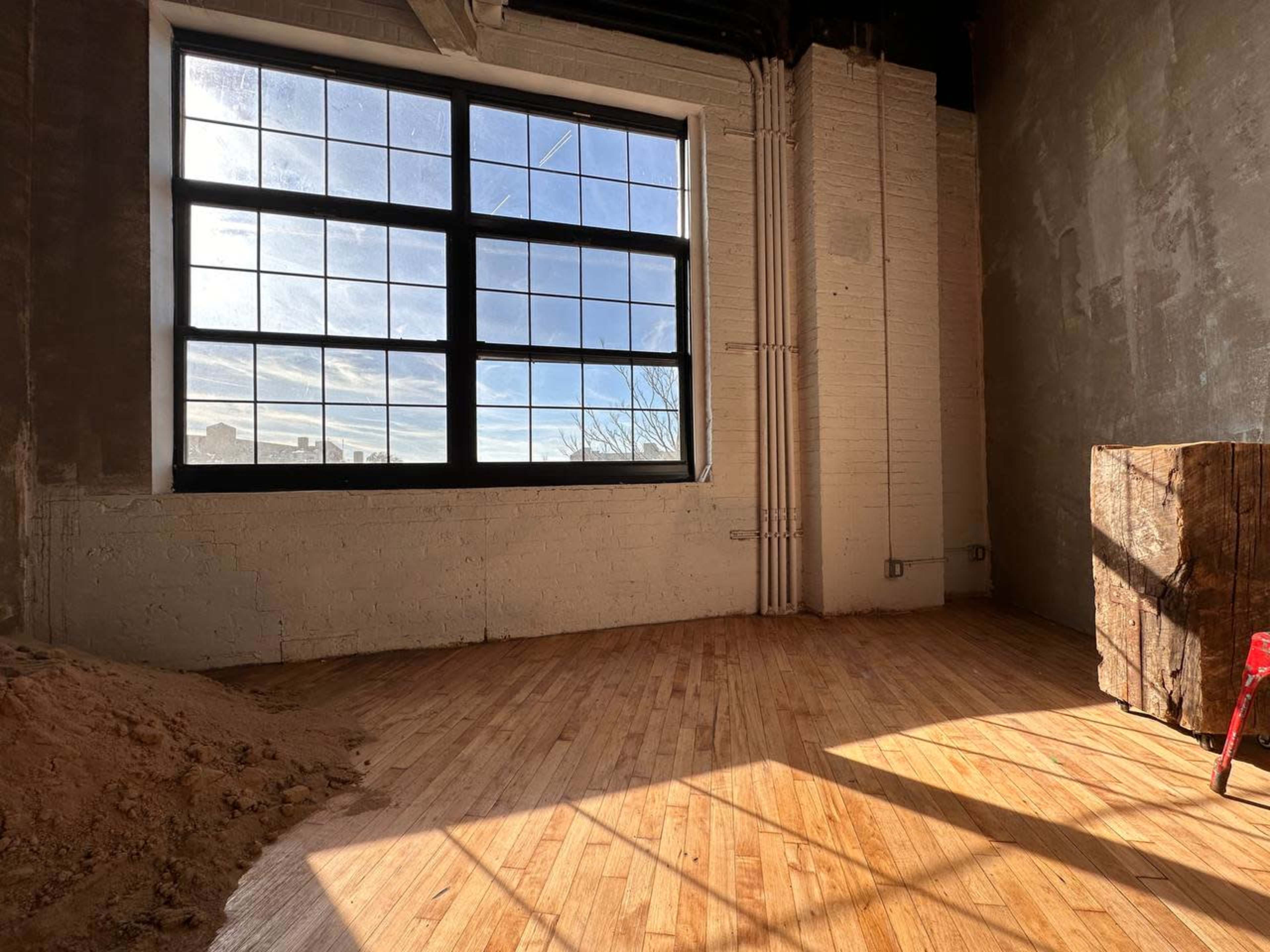 A large industrial window provides natural light to a room with wooden flooring and a partially finished wall.